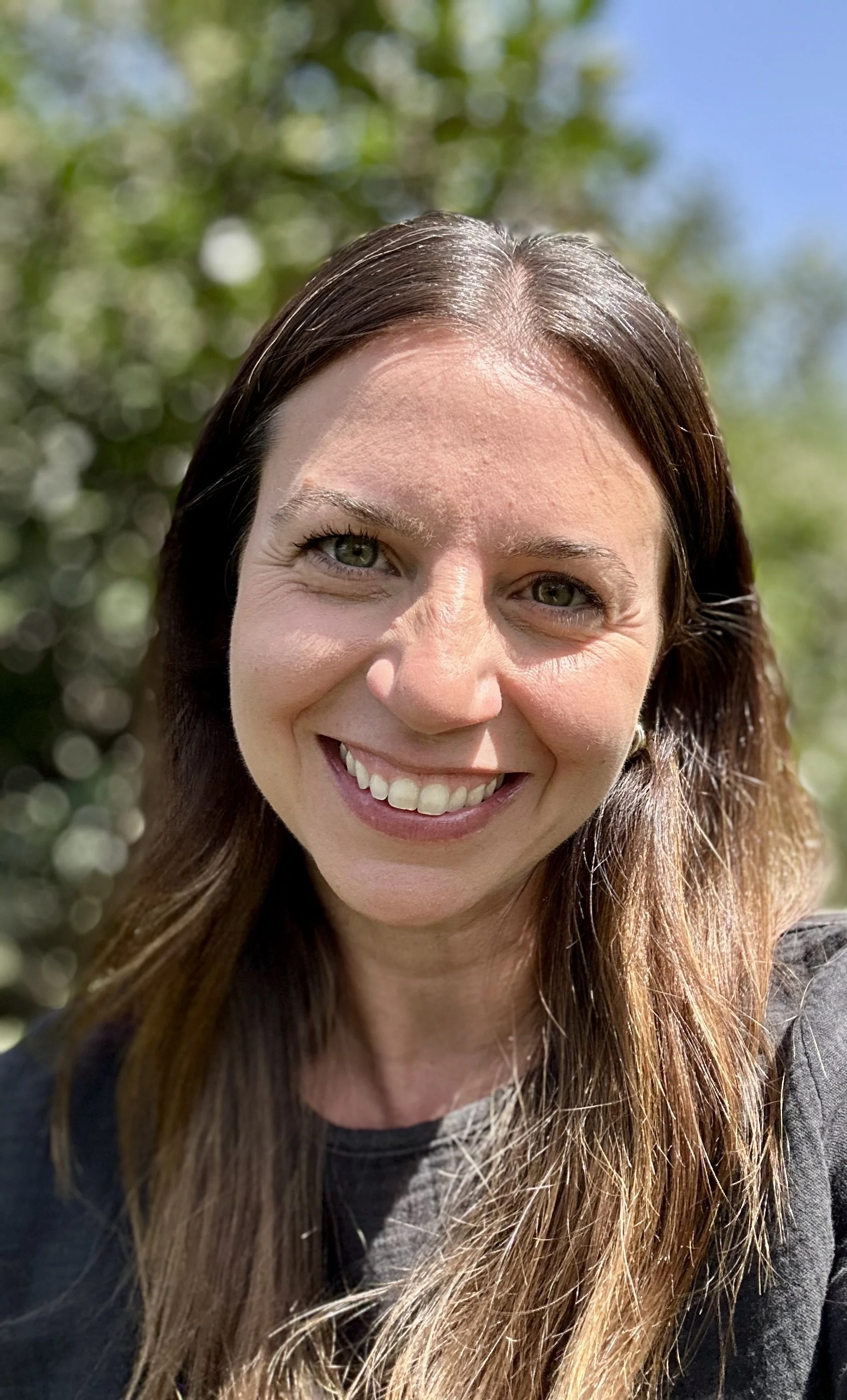 A woman, Kristen Vernace CFP CDFA, with long brown hair smiling outdoors with blurred trees in the background.