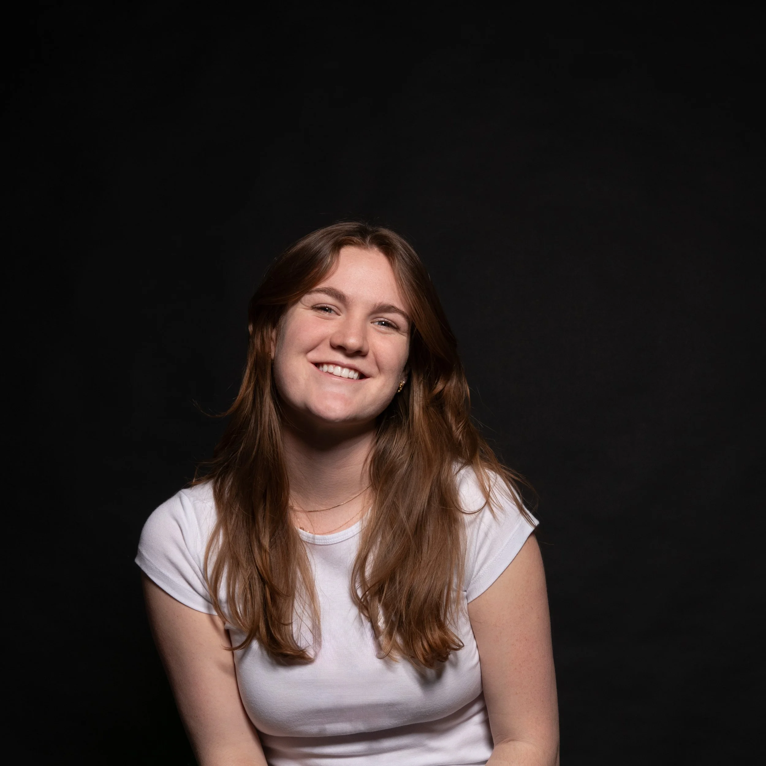 A young woman with long brown hair smiling against a black background.
