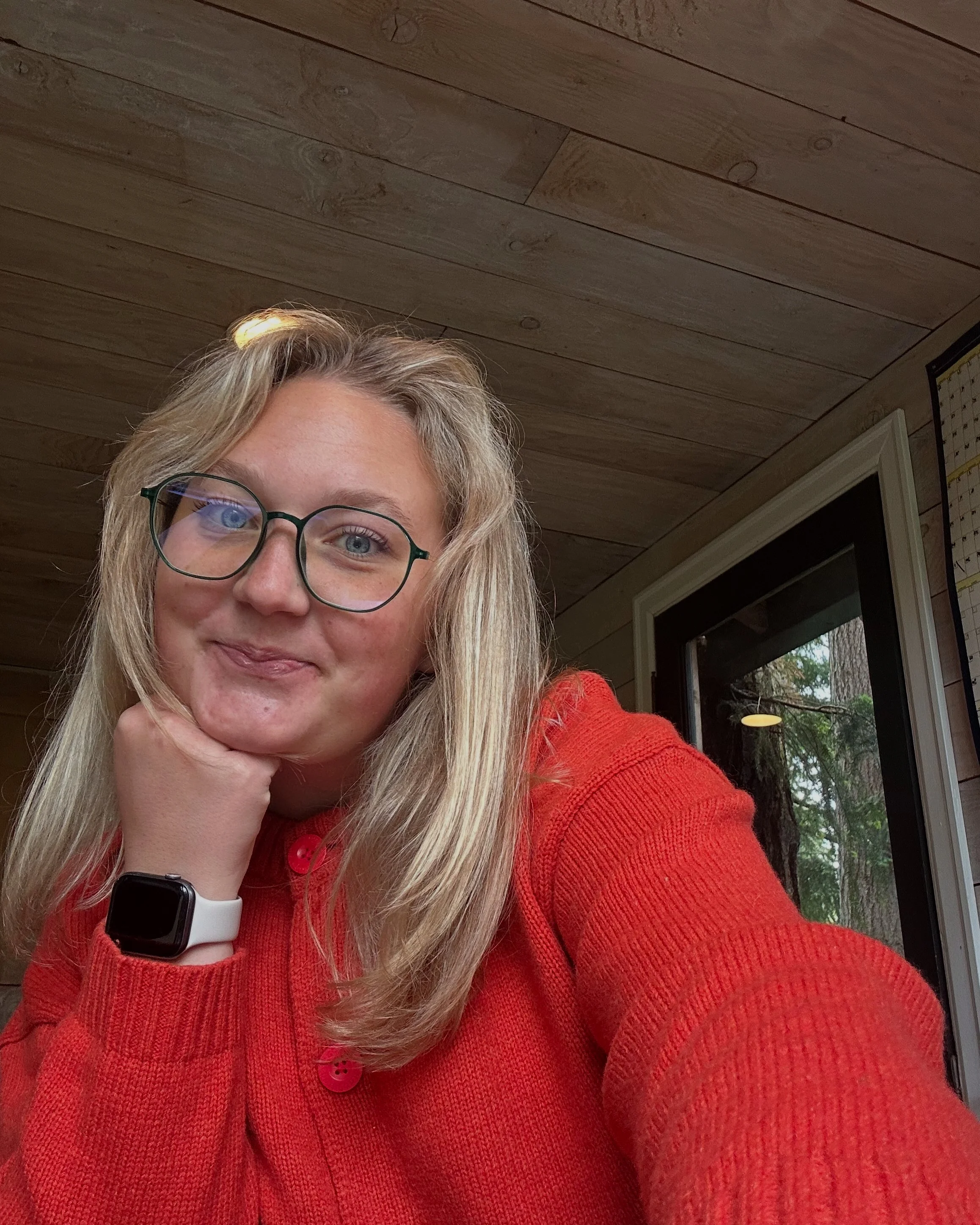 A young woman with blonde hair, glasses, and a smartwatch, wearing a red sweater, resting her chin on her hand and smiling while taking a selfie indoors with a wood-paneled ceiling and a window showing trees in the background.