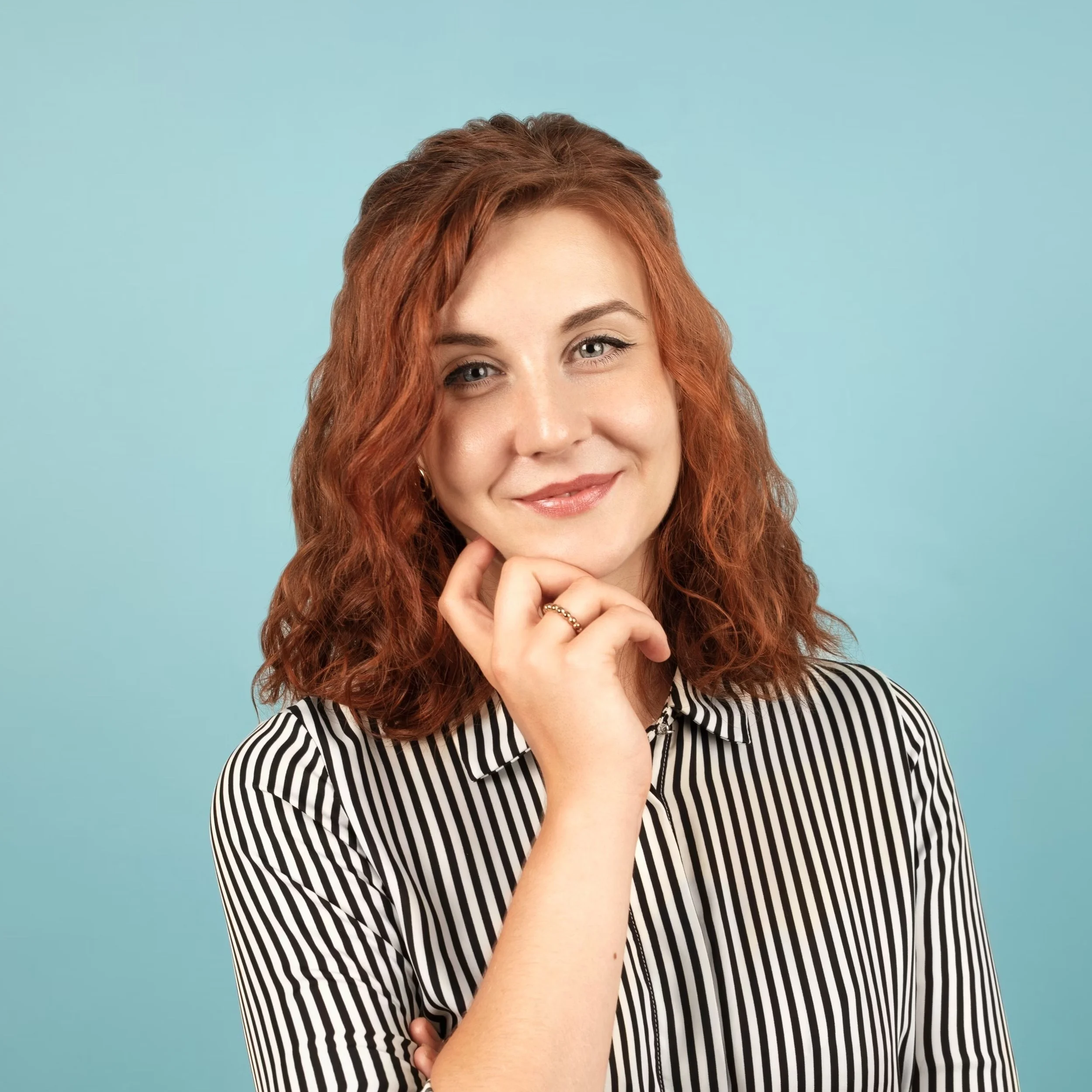 Person with red curly hair, wearing a striped shirt, posing against a blue background.