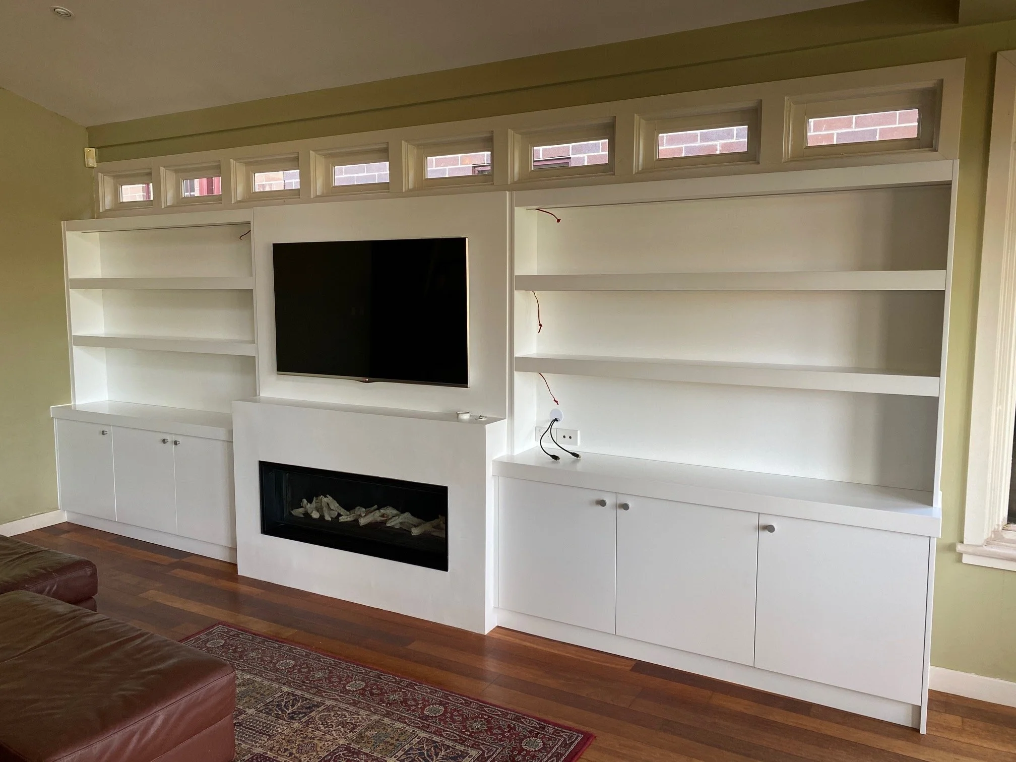 Living room with white entertainment center, three shelves on each side, a mounted flat-screen TV, built-in fireplace below, and a row of high small windows above. Part of a maroon sofa and a patterned area rug are visible.