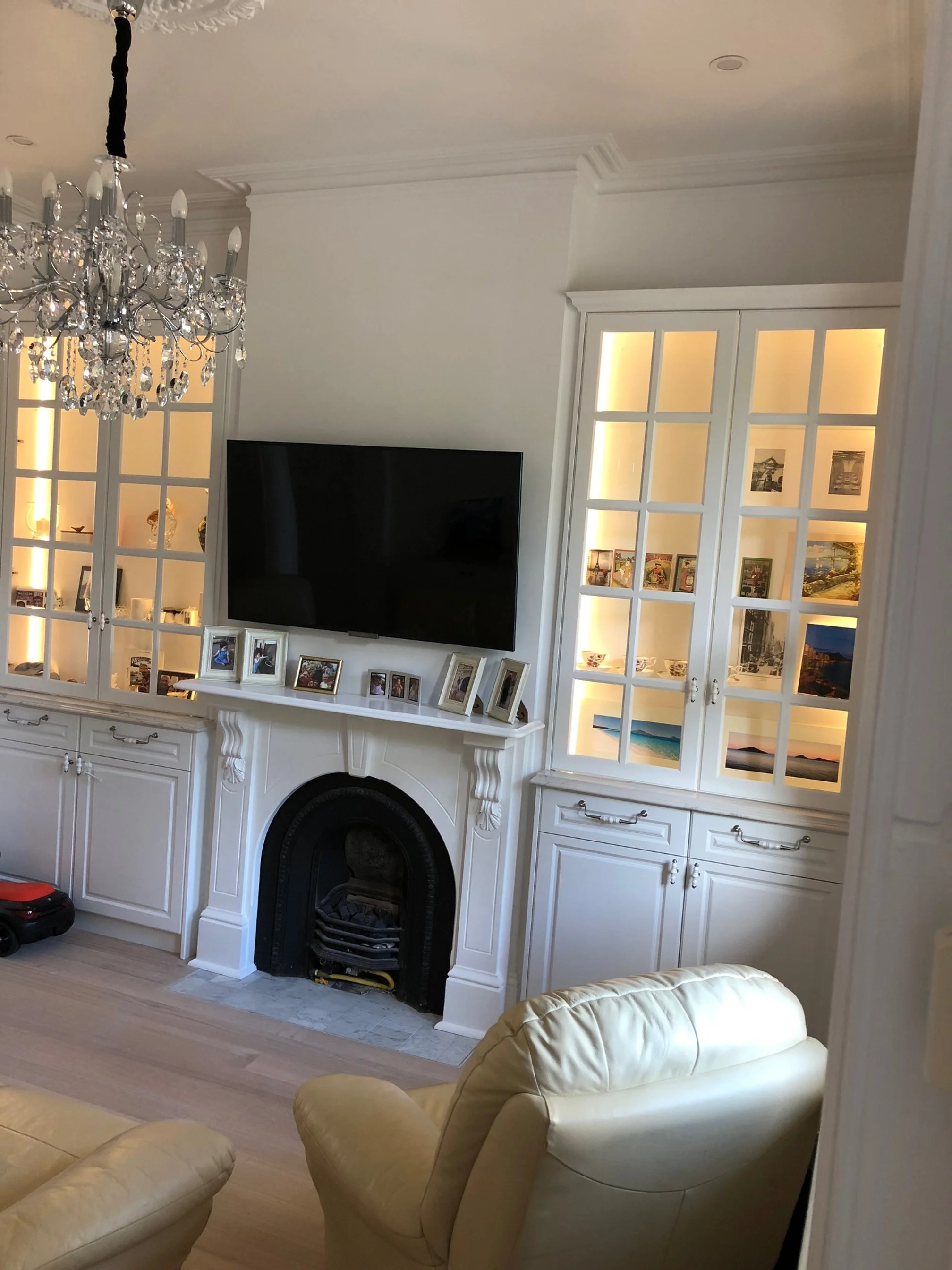 Living room with a fireplace, a large flat-screen TV, and built-in display cabinets illuminated from within. There is a chandelier hanging from the ceiling and a cream-colored sofa in the foreground.