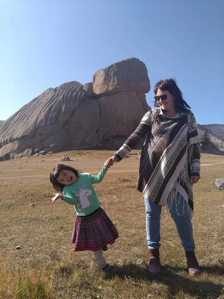 Amber De Vogt and child holding hands near a large rock formation outdoors.
