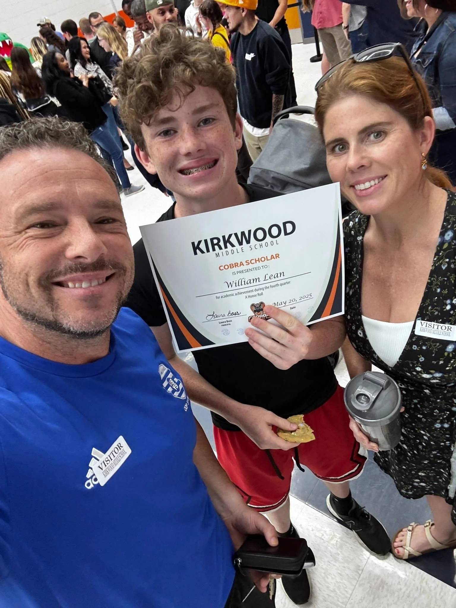 A family celebrating a middle school graduation, holding a diploma for William Lean, with many students and staff in the background.