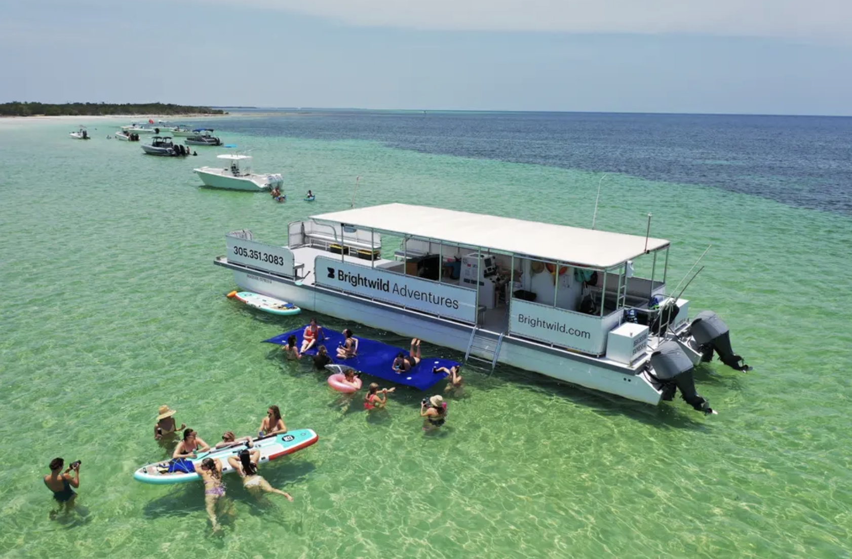 A boat with a person standing on it, looking at their phone, on clear turquoise water near a shoreline.