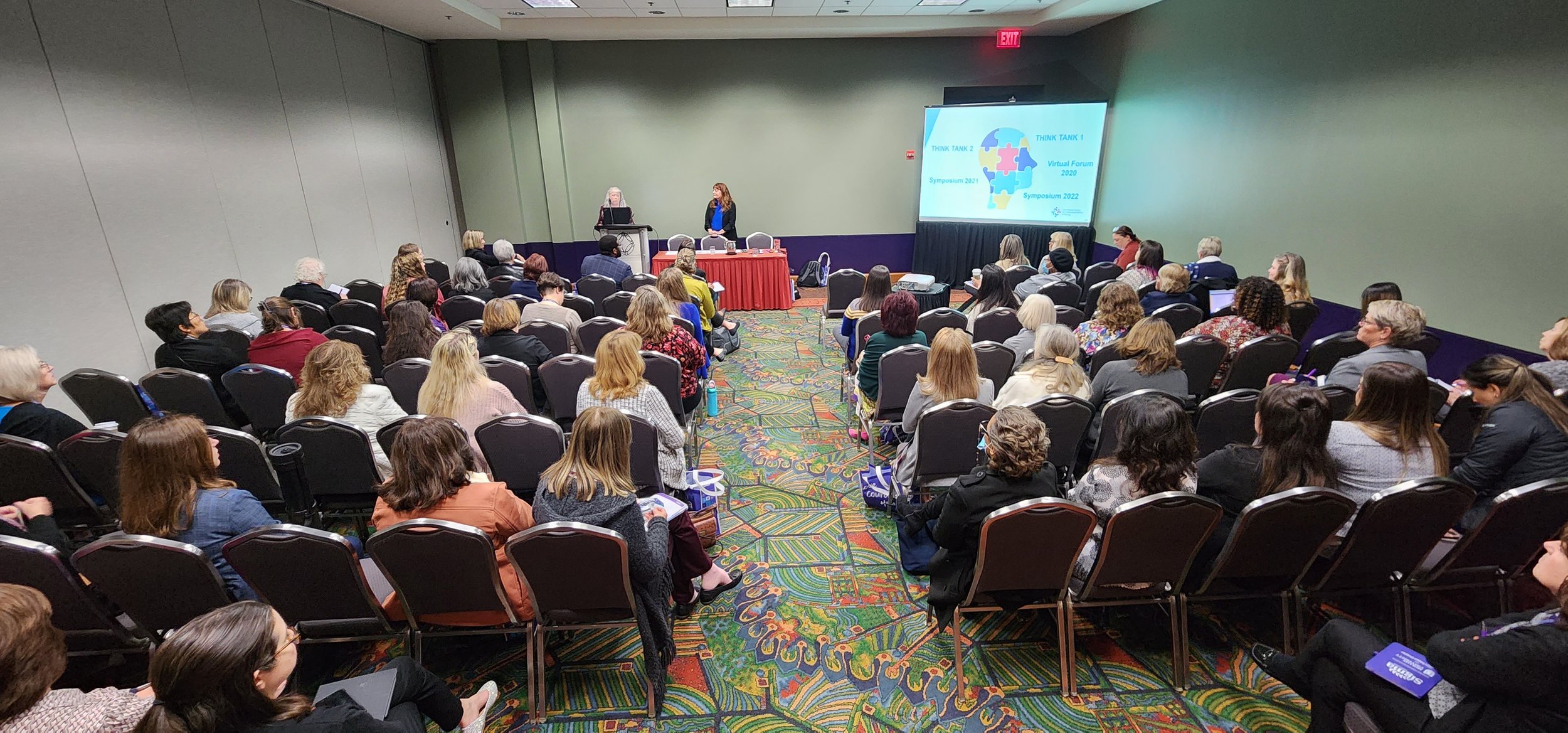 Conference room with attendees listening to a presentation, two speakers at the front, and a projector screen displaying event information.