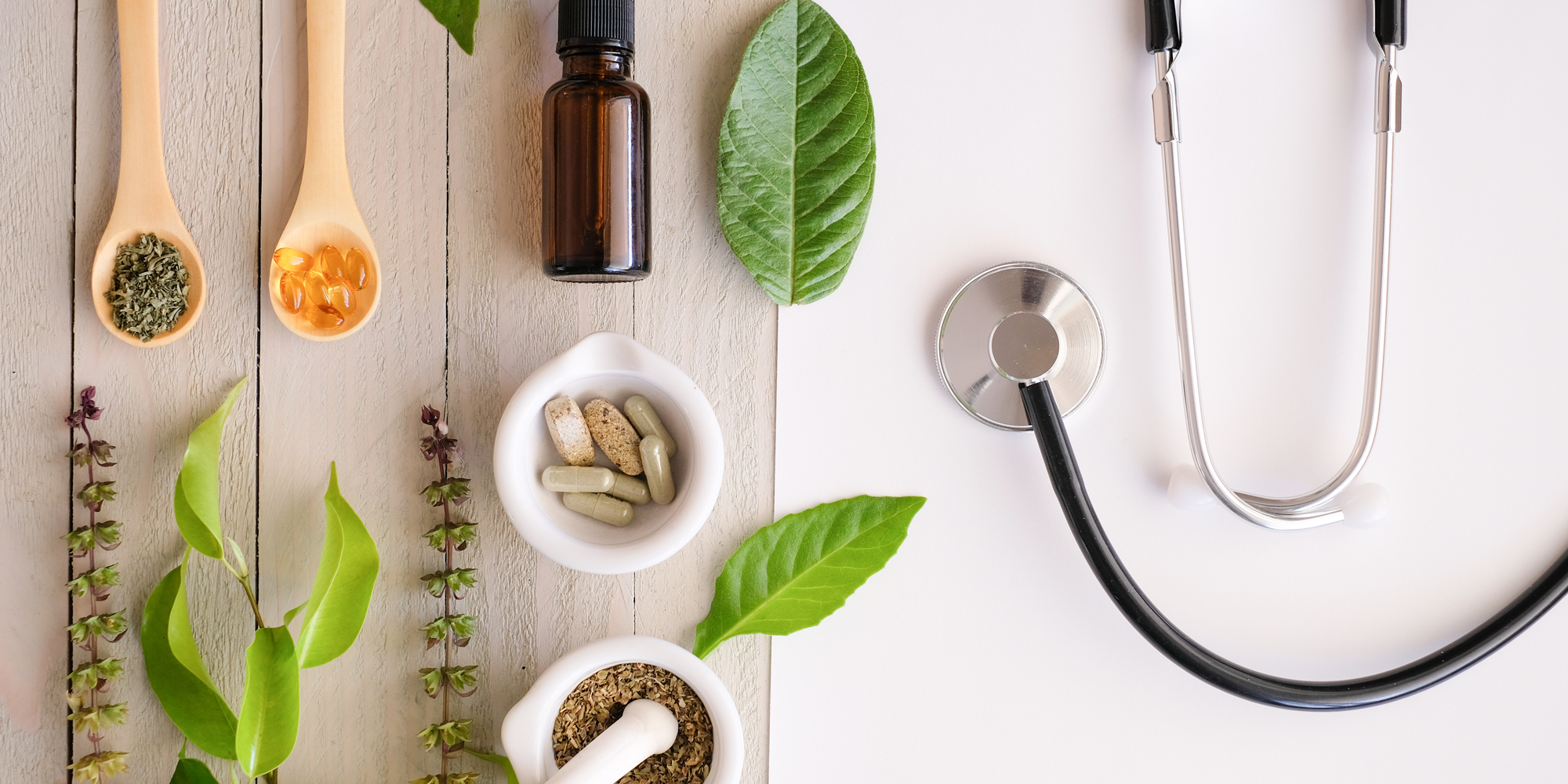 An arrangement of natural health products including dry herbs, capsules, leaves, and a stethoscope on a wooden and white background.