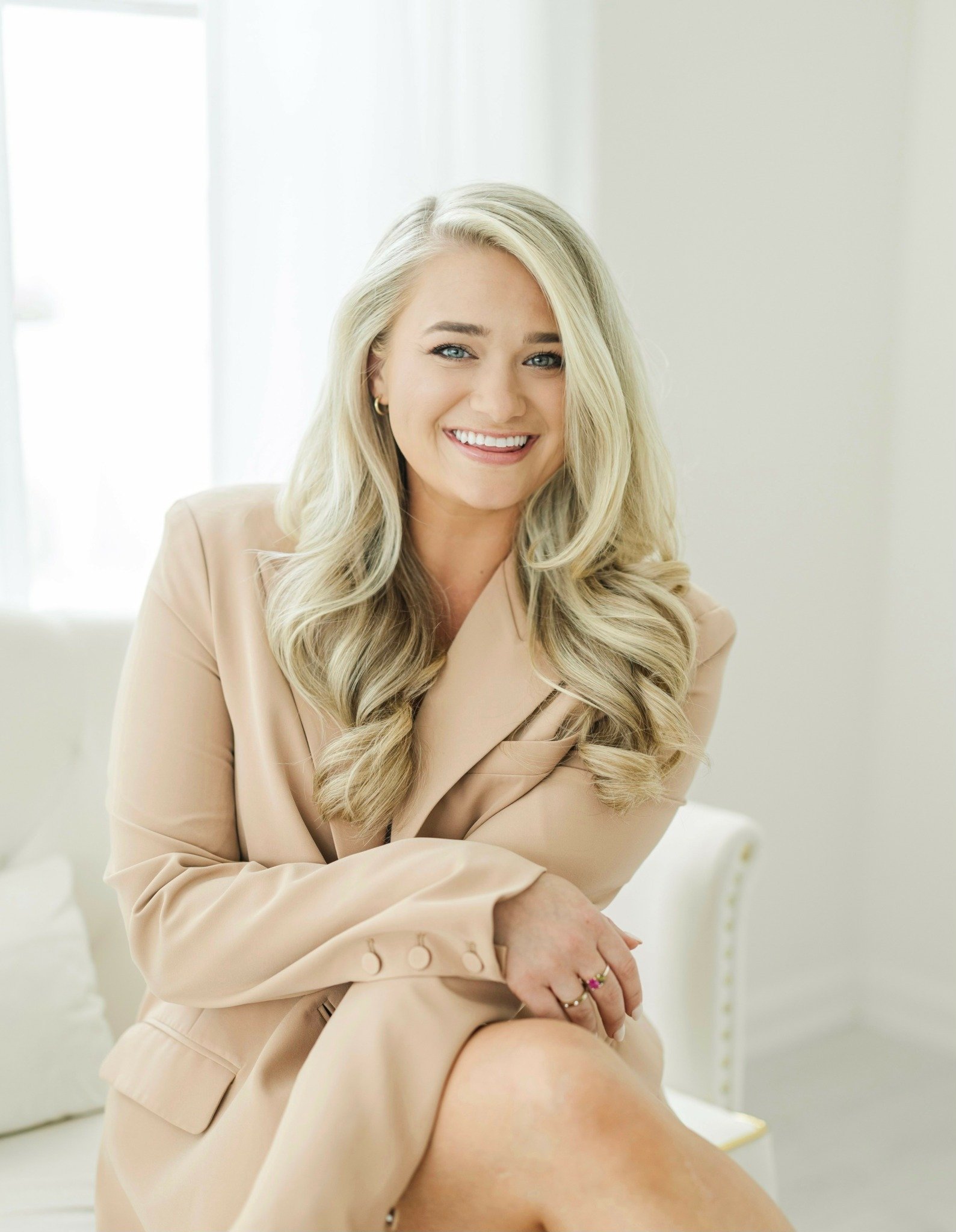 A smiling blonde woman with long, wavy hair, wearing a beige blazer, sitting on a white couch in a bright, softly lit room.