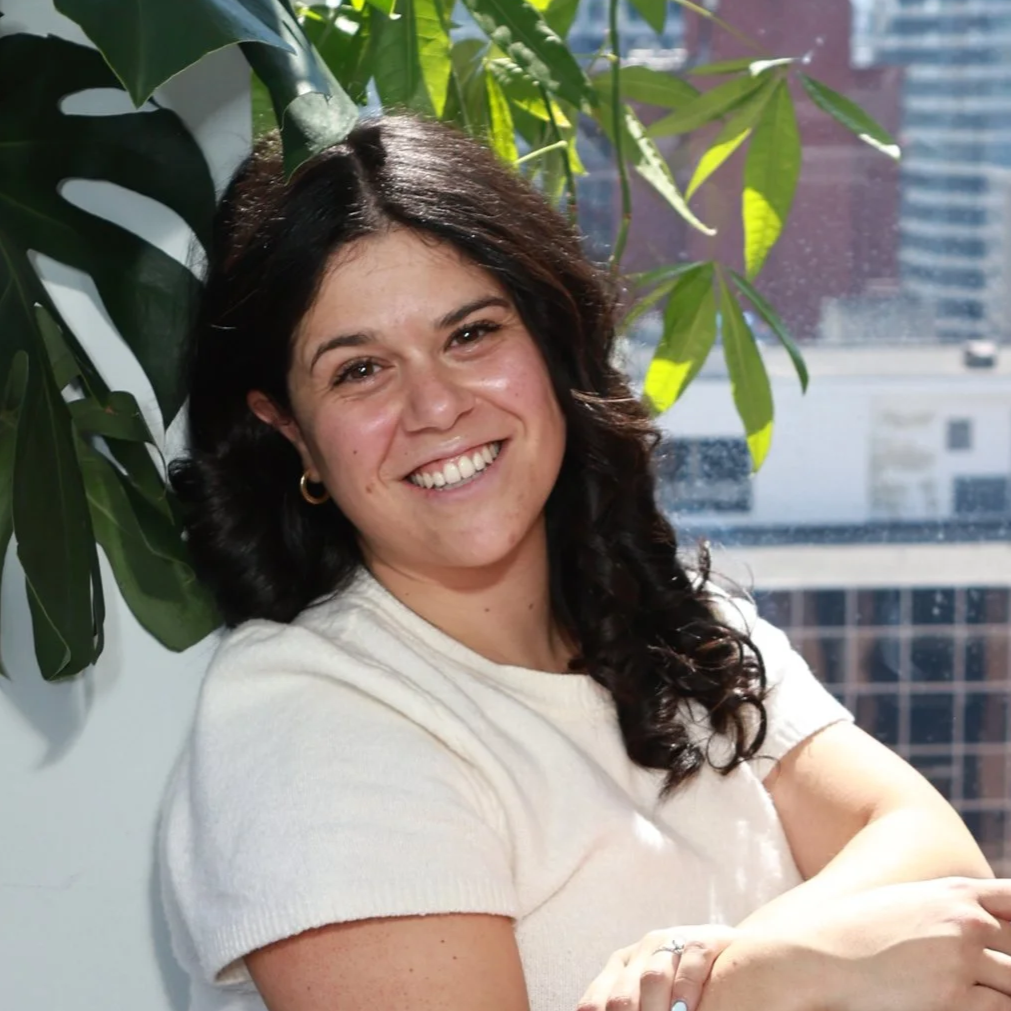 Woman with brown hair facing the camera smiling. Plant in background and window with buildings in it.