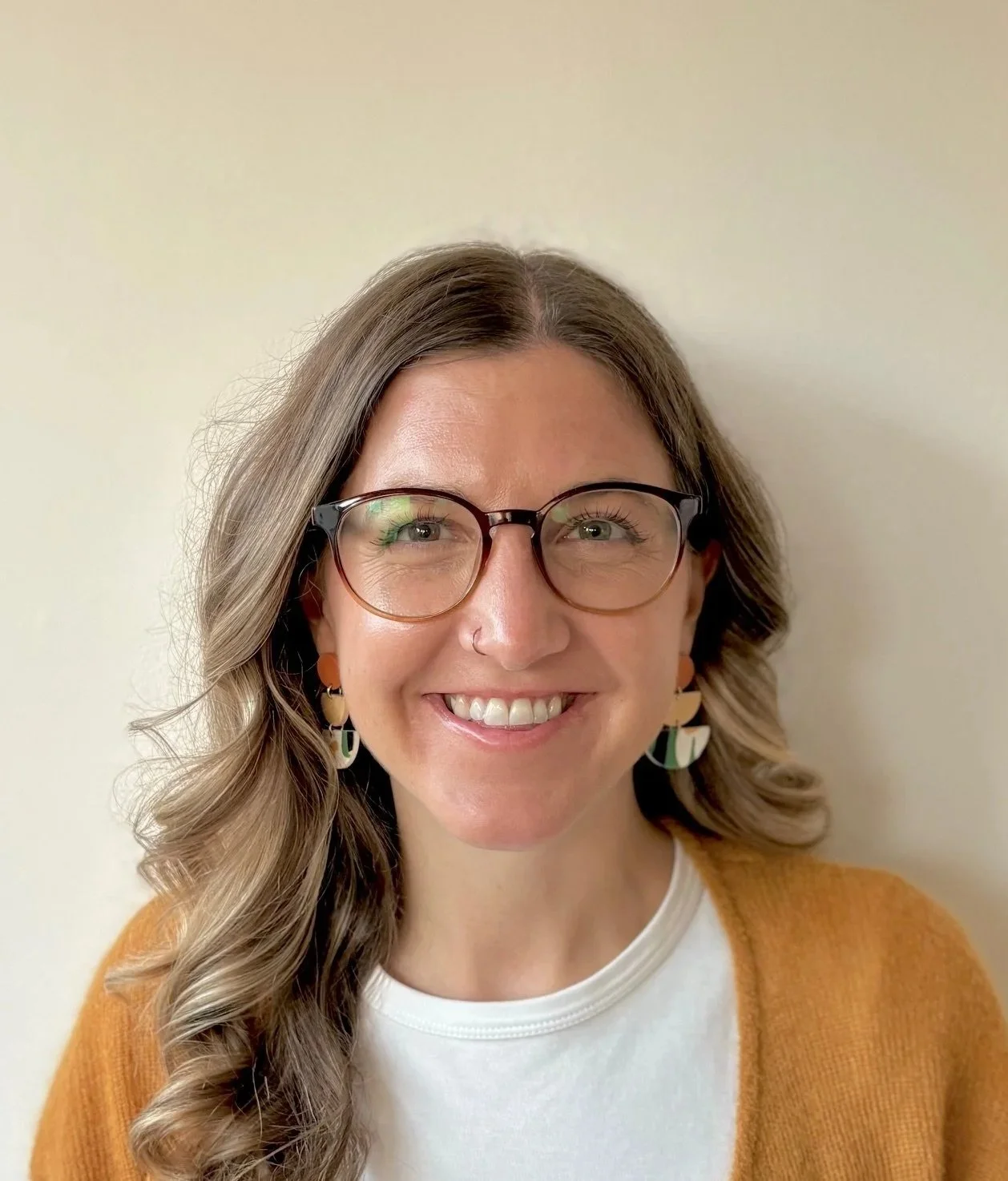 Woman with blonde hair, wearing glasses, and smiling. Woman is looking directly at the camera. Neutral wall as the background. 