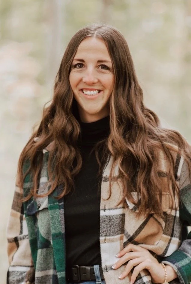 A woman in flannel and brown hair facing the camera with her hand on her hips