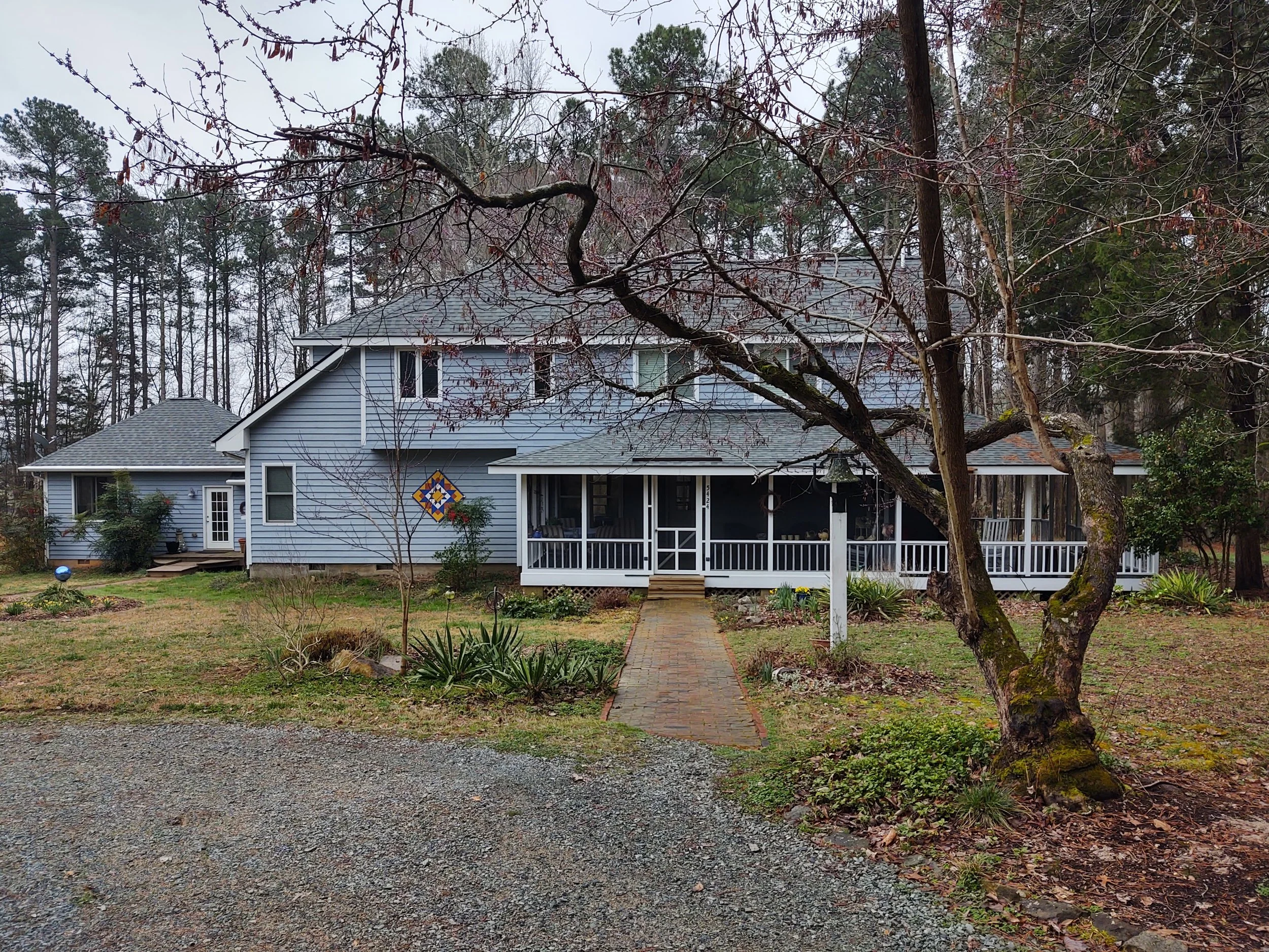 A two-story light blue house with a screened porch, surrounded by a yard with trees and bushes, cloudy sky, and a gravel driveway.