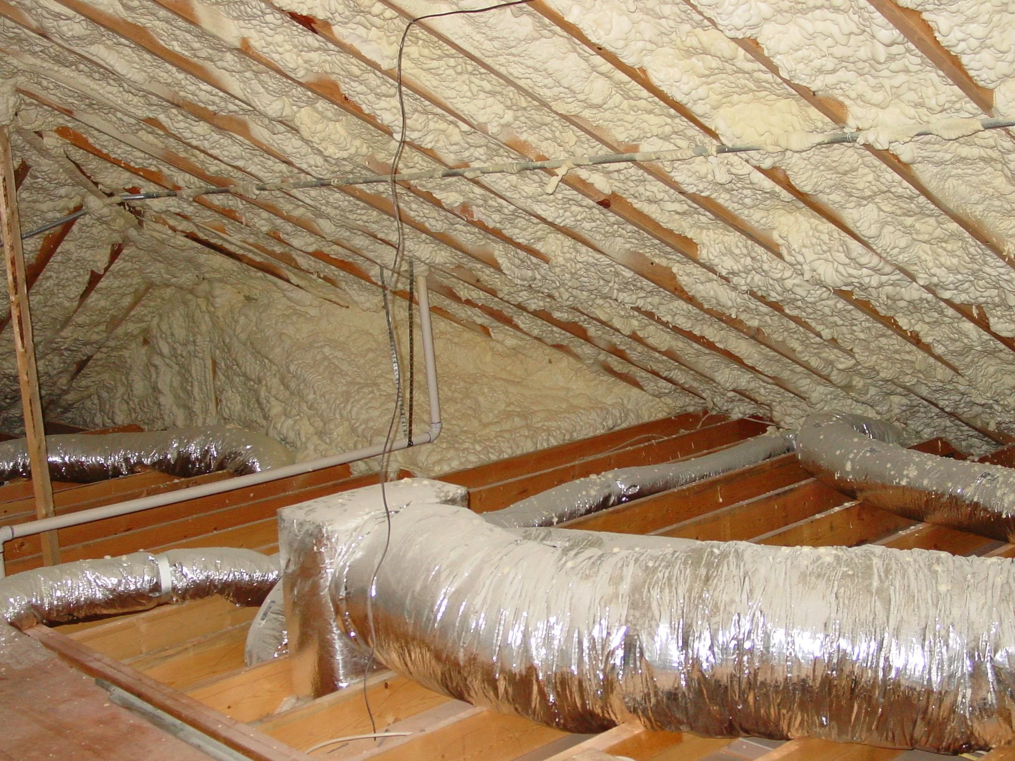 Insulated attic space showing foam spray insulation, ductwork, wiring, and wooden beams.