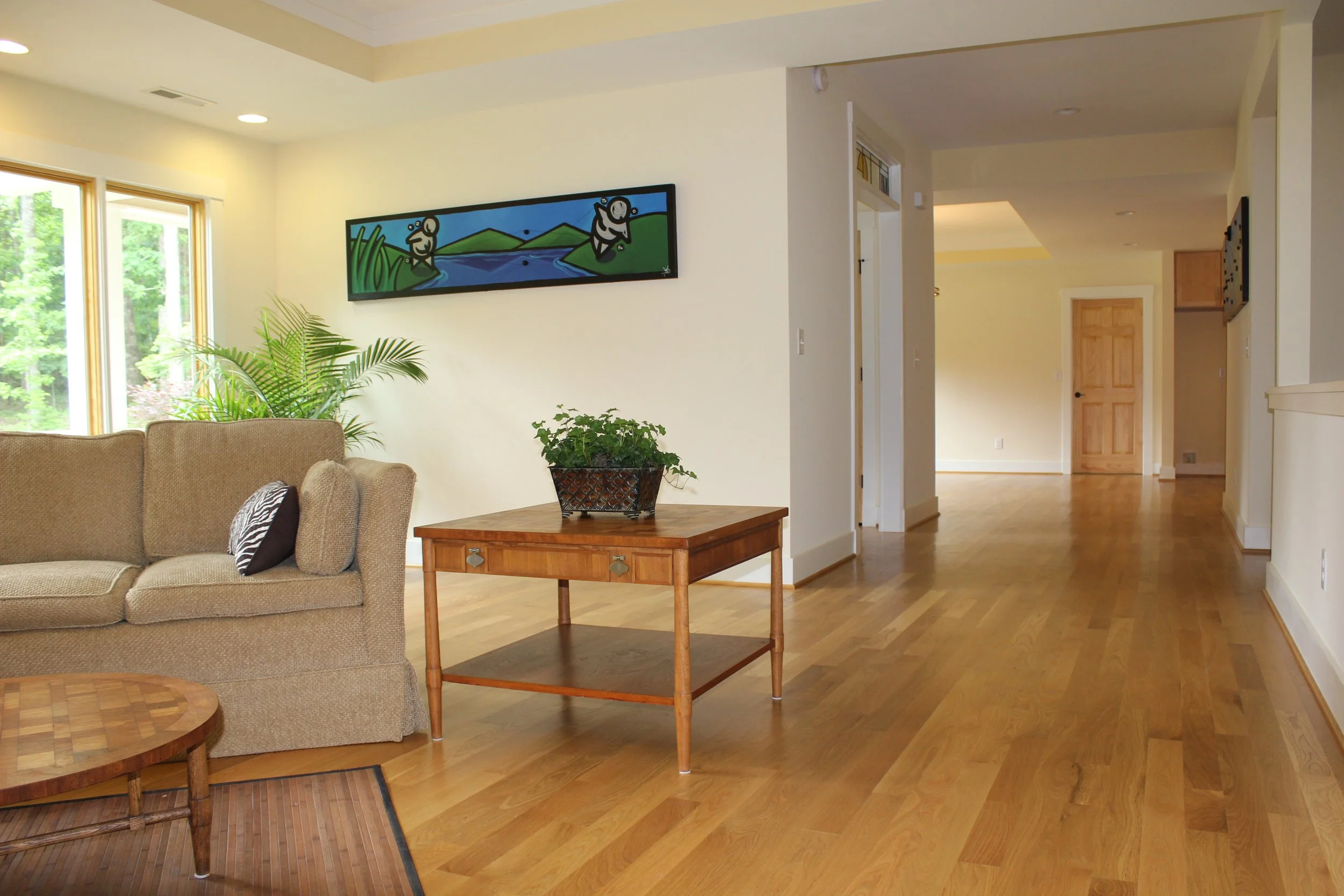 Living room with beige sofa, green plants, wooden coffee table, and hallway with wooden floors and doors.
