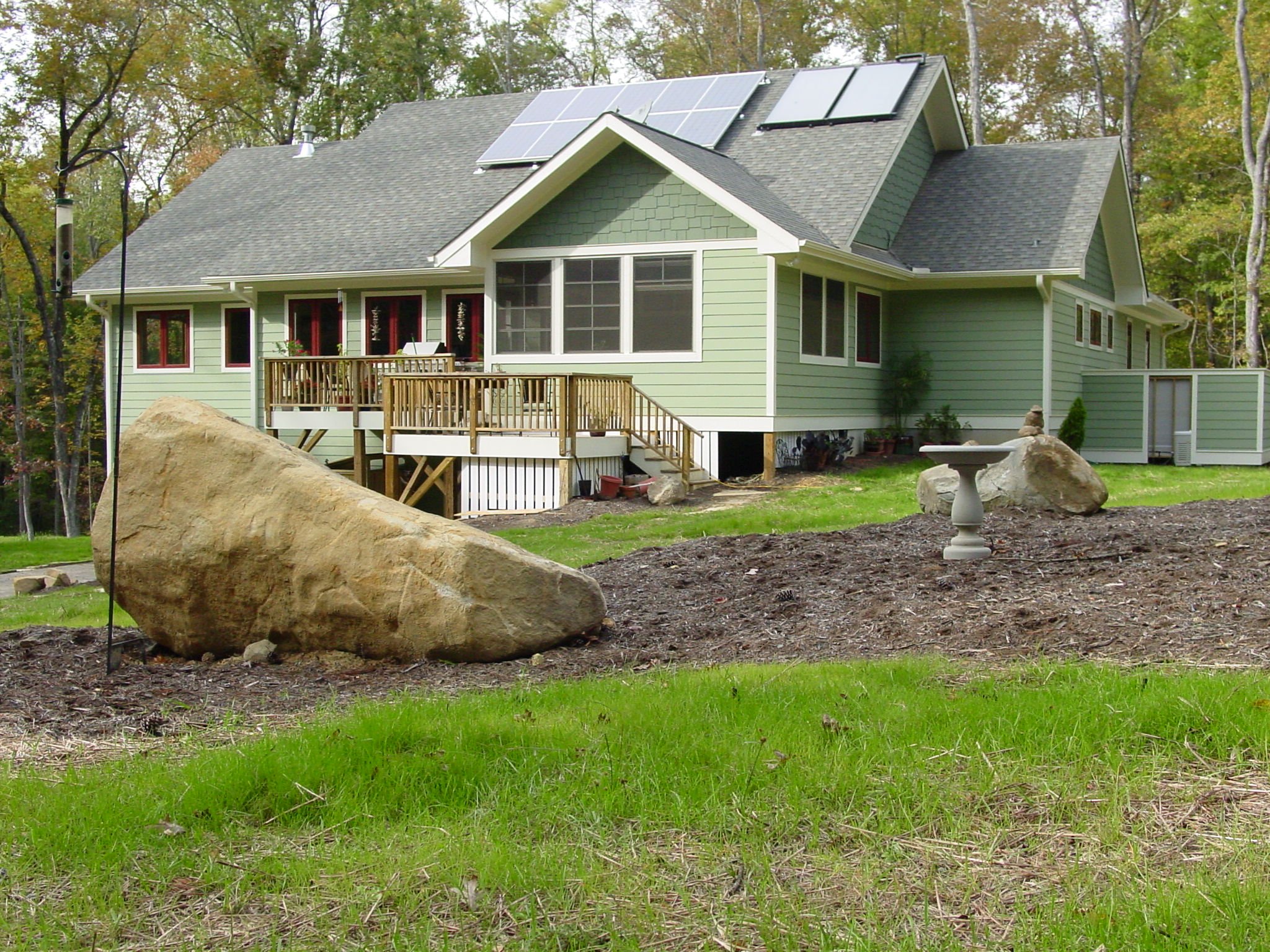 Green house with solar panels on the roof, surrounded by trees, with a wooden deck, rocks, and a birdbath in the yard.