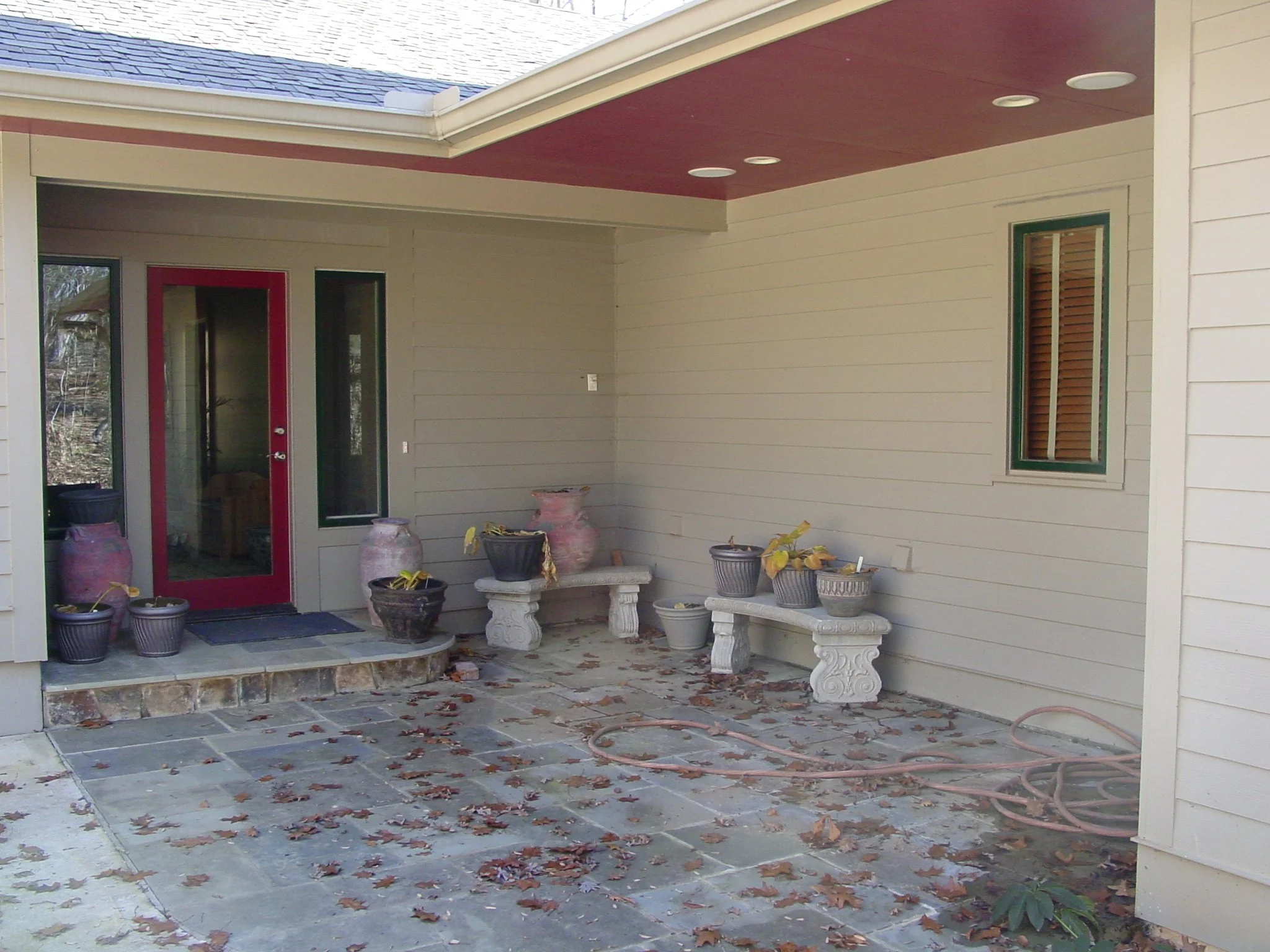 Covered patio area with potted plants on benches, fallen leaves on the ground, and a red door with glass panel.