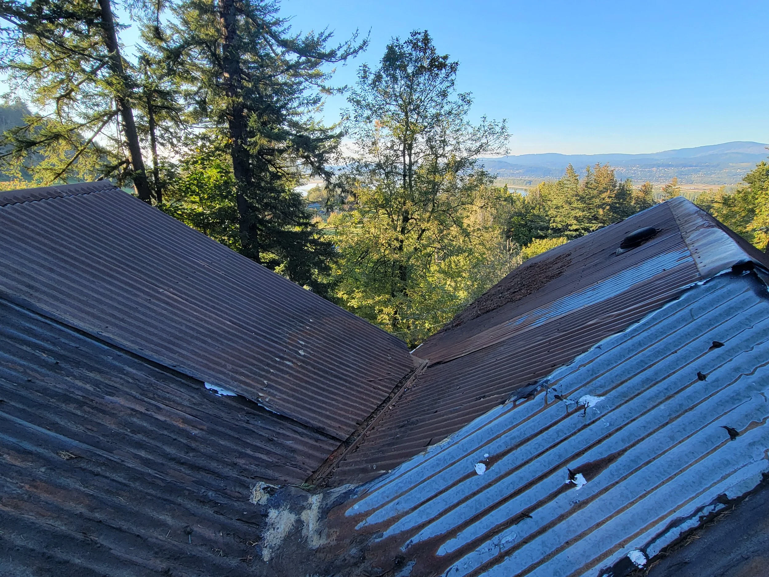 Close-up view of a metal roof with two interconnected sections, surrounded by trees and a distant view of hills and sky in the background.