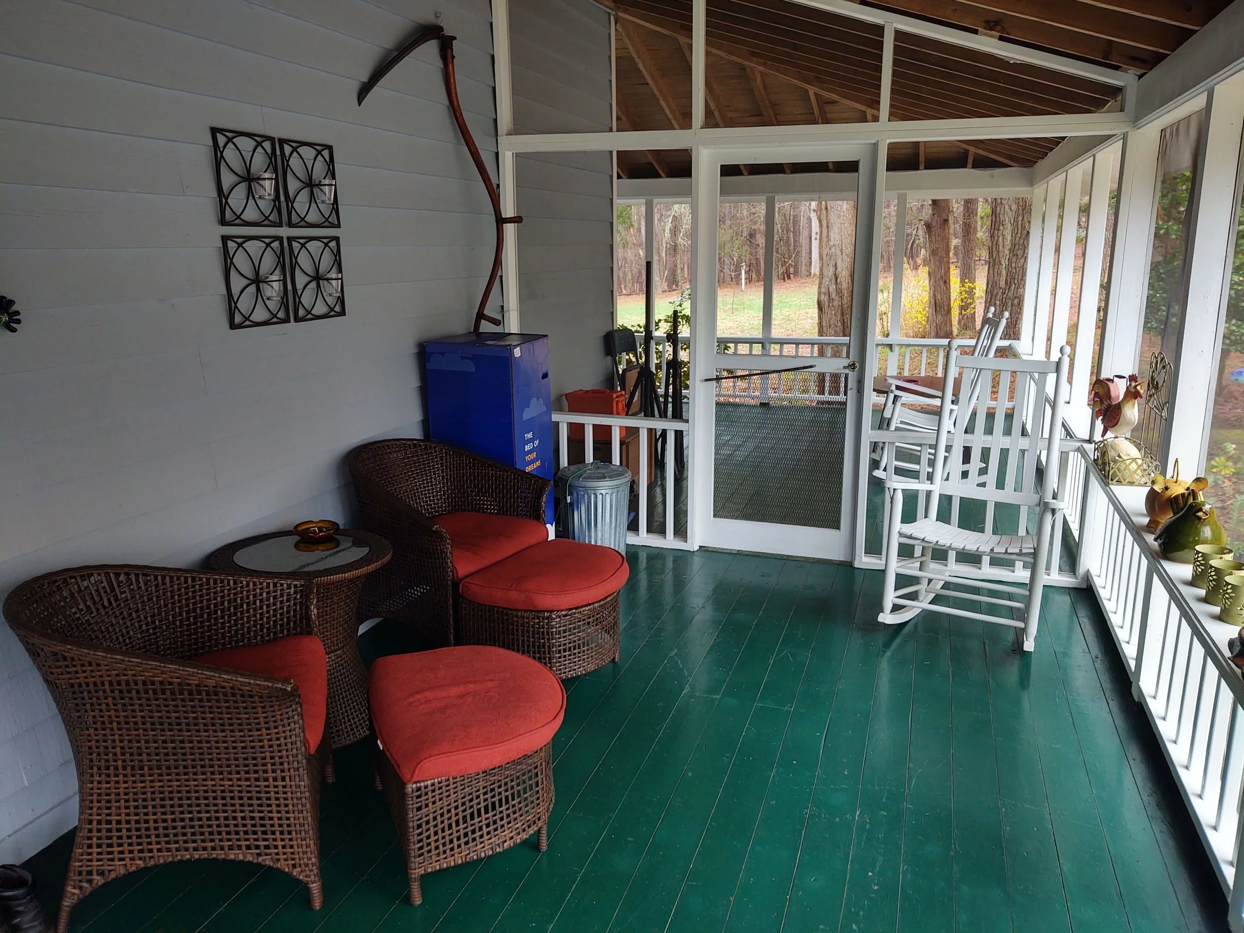 Enclosed porch with wicker chairs, a small table with a ashtray, a blue refrigerator, and decorative items on a windowsill. The porch has white railings and a view of trees outside.