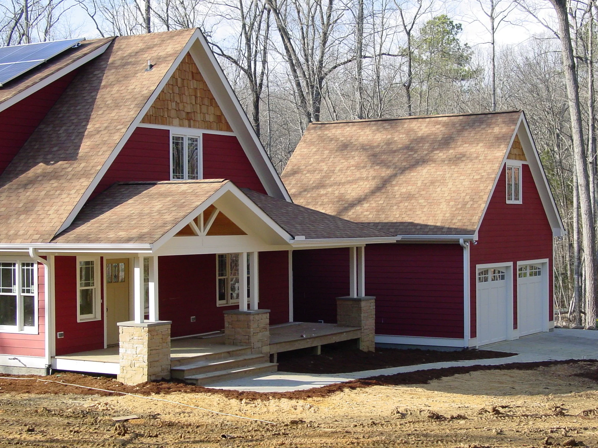 Newly built red house with white trim, multiple gable roofs with shingles, front porch with steps and stone pillars, and attached garage, surrounded by bare trees.