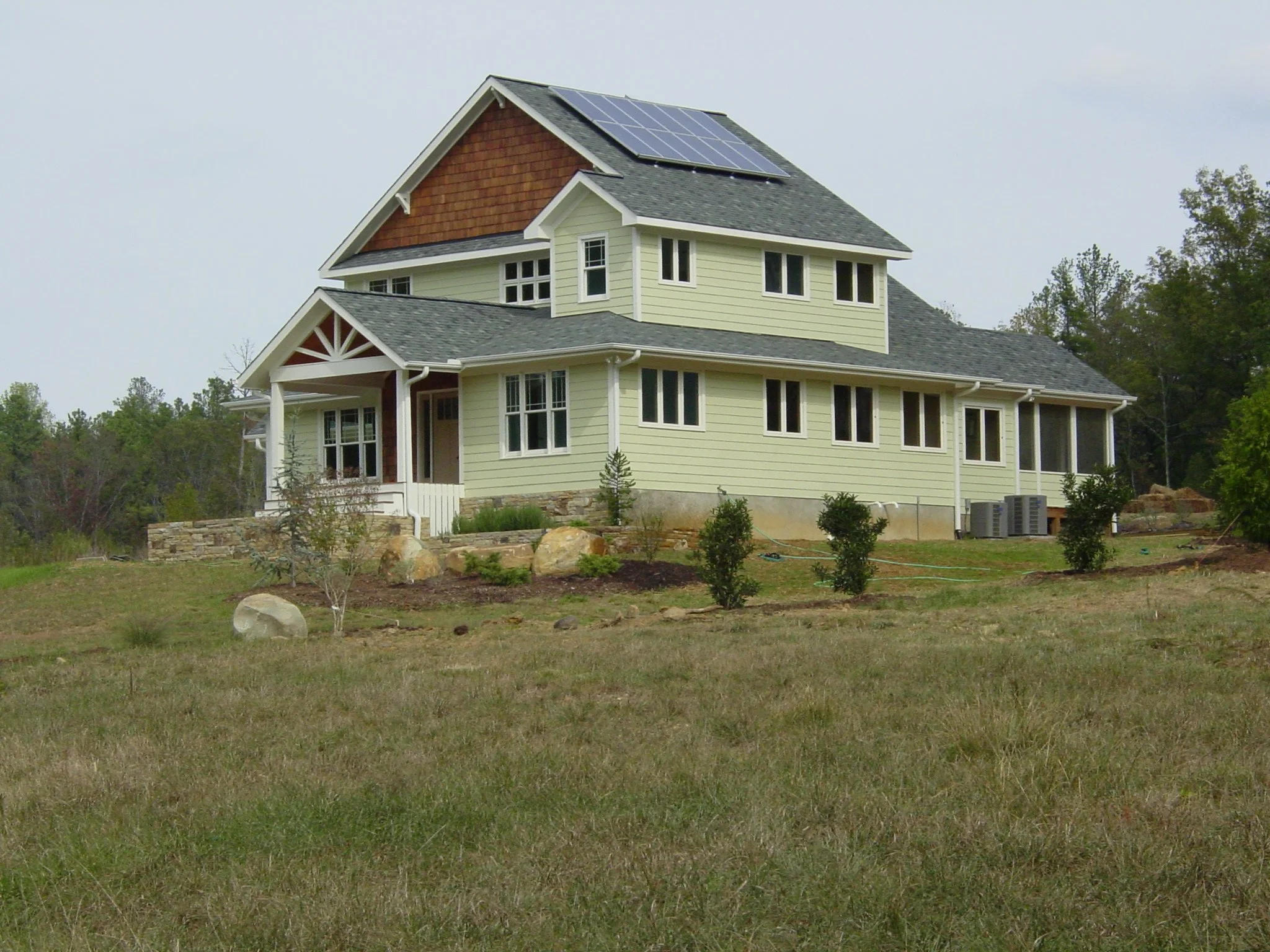 A green two-story house with solar panels on the roof, surrounded by grass and small bushes, with trees in the background.