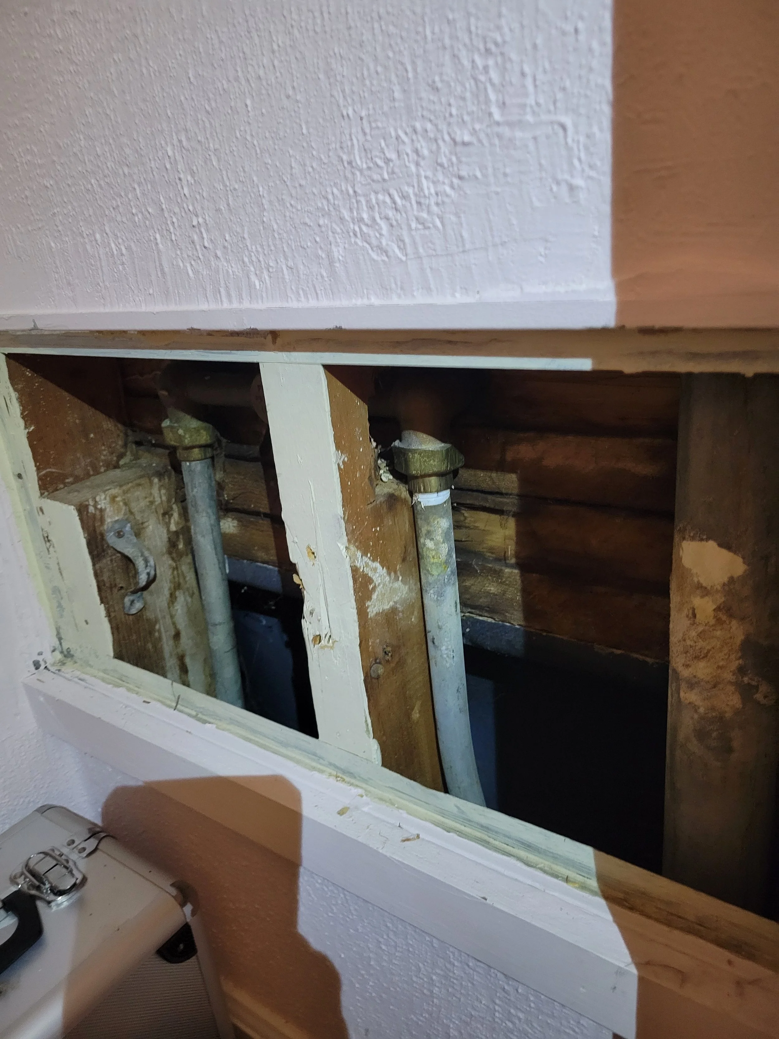 View of a wall with an open rectangular hole revealing lead plumbing pipes and wooden framing inside the wall