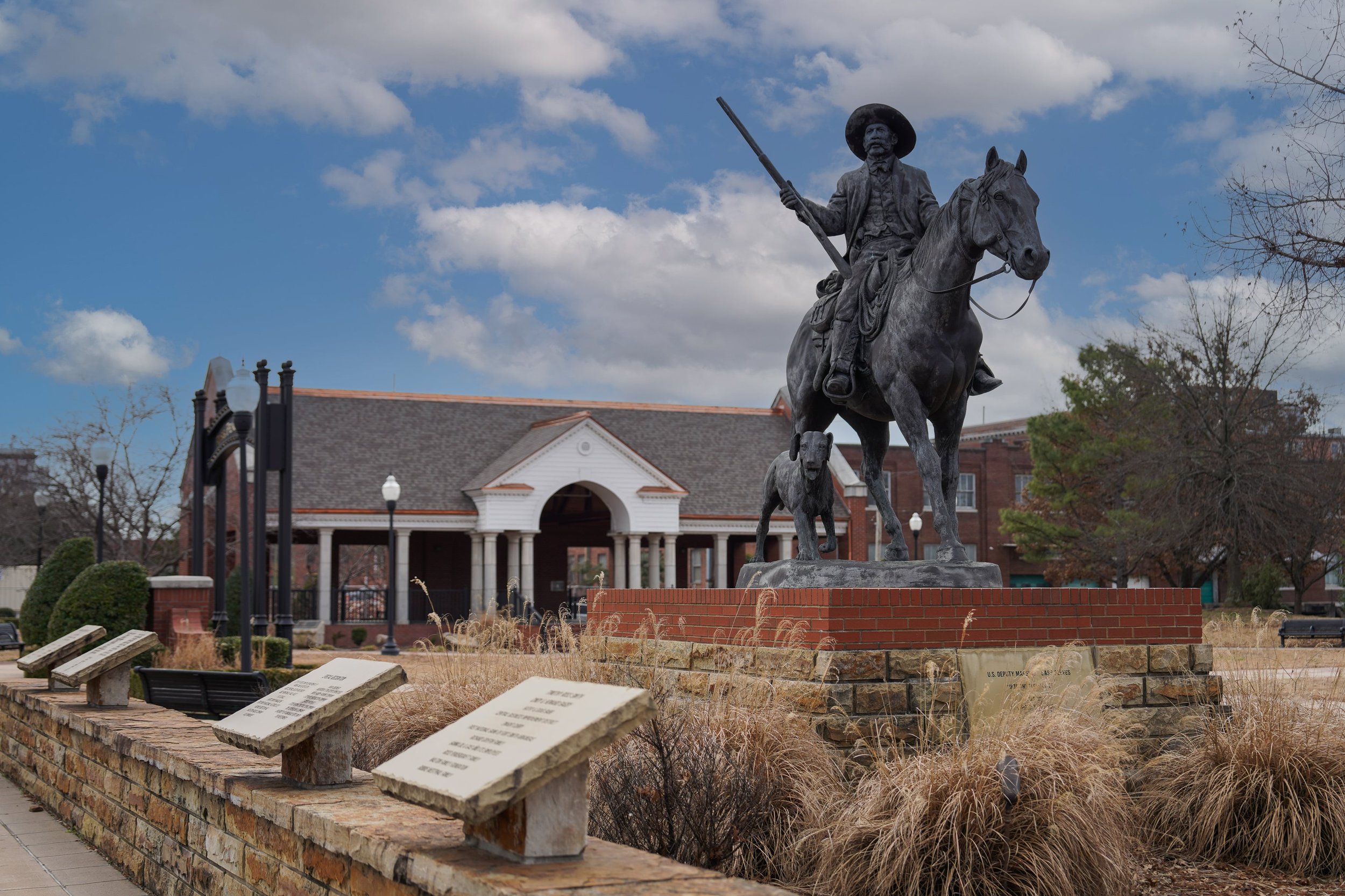 Outdoor statue of a man on horseback holding a rifle, accompanied by a dog, with a historical building in the background and information plaques in the foreground.