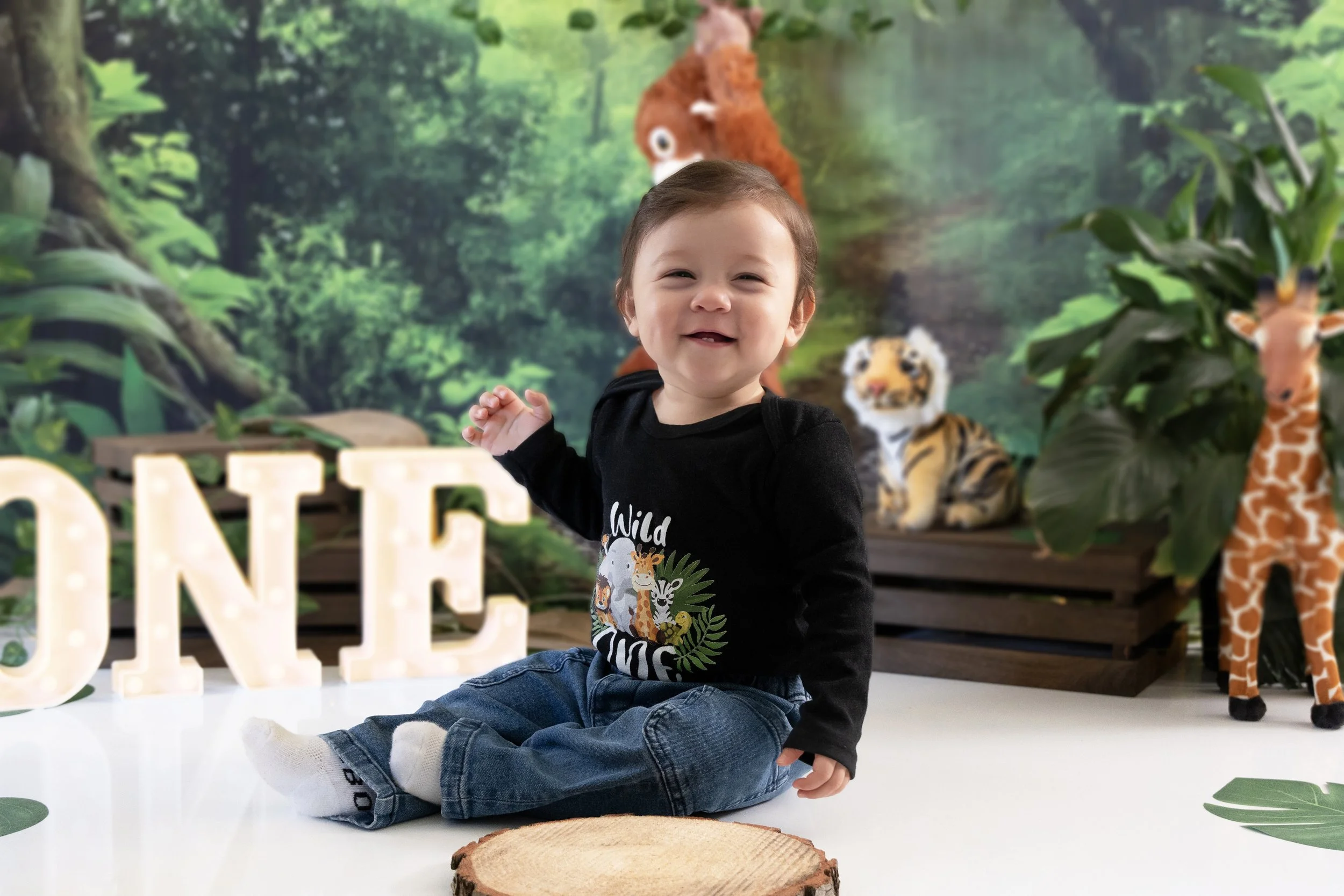 A young child celebrating their first birthday with a cake, surrounded by balloons, a large gold number one balloon, and colorful bunting. The child is holding a teddy bear with frosting on its face, and there is a mint green decorated cake in front.