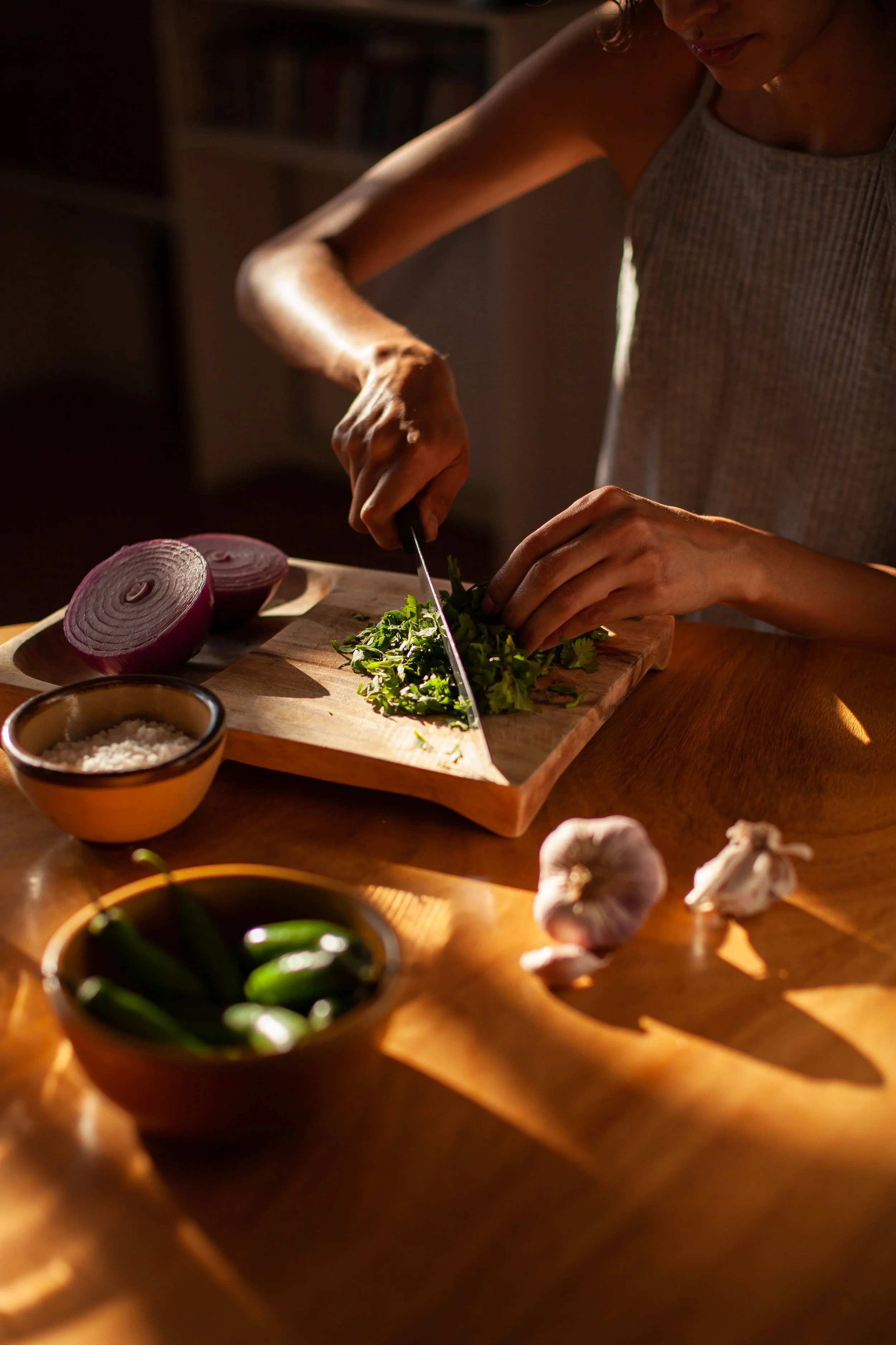 A person chopping fresh herbs on a wooden cutting board with red onion and other ingredients nearby, in a warmly lit kitchen.