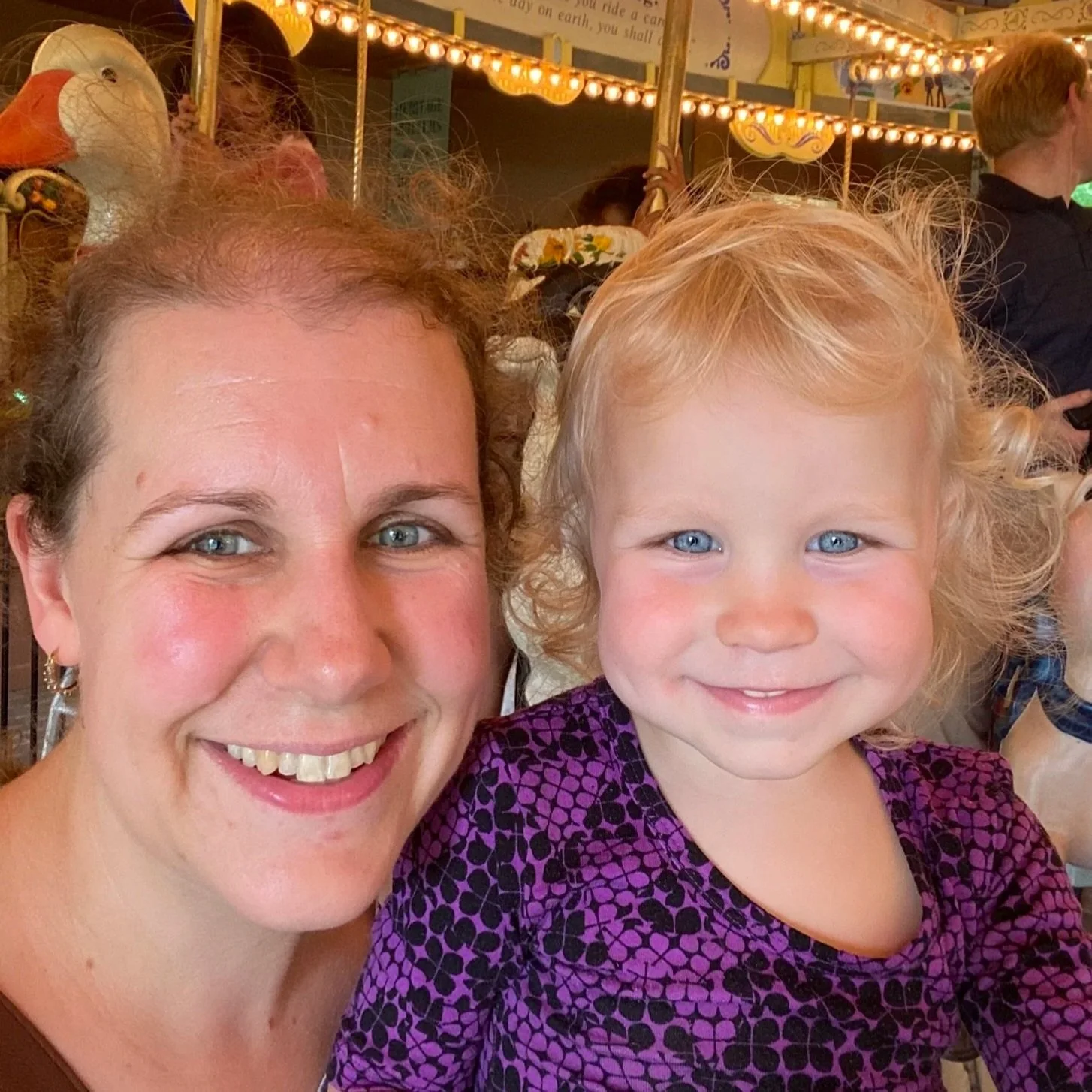 A smiling woman and a young girl with curly blonde hair, both with blue eyes, close together in a fun amusement park or carnival setting with lights and rides in the background.