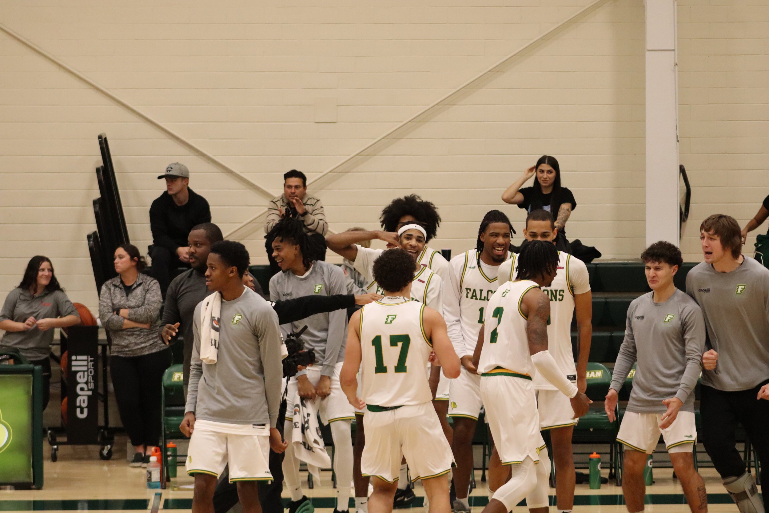 A group of basketball players and spectators in an indoor gymnasium. The players are wearing white jerseys with green accents and the word 'Falcons' printed on them, and some have tattoos and headbands. Some individuals appear to be celebrating or ch