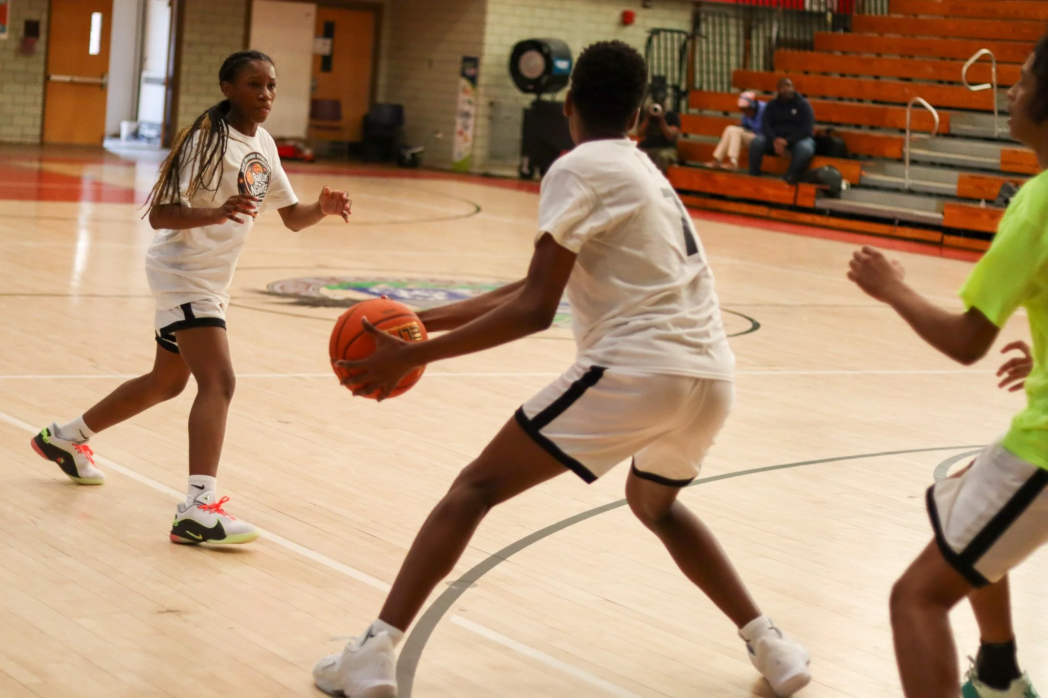 Children playing basketball on an indoor court, with bleachers and spectators in the background.