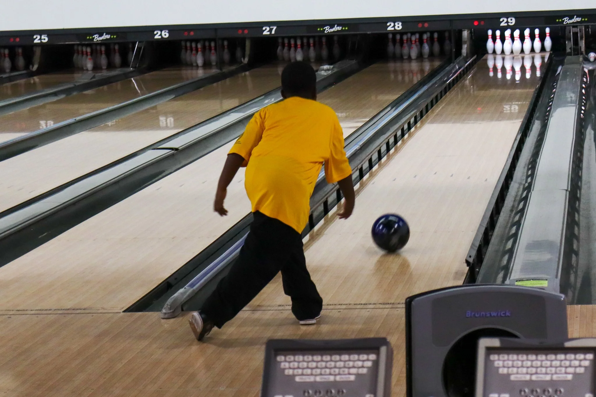 A person in a yellow shirt is bowling on a lane at a bowling alley, with bowling pins at the end and a ball in motion.