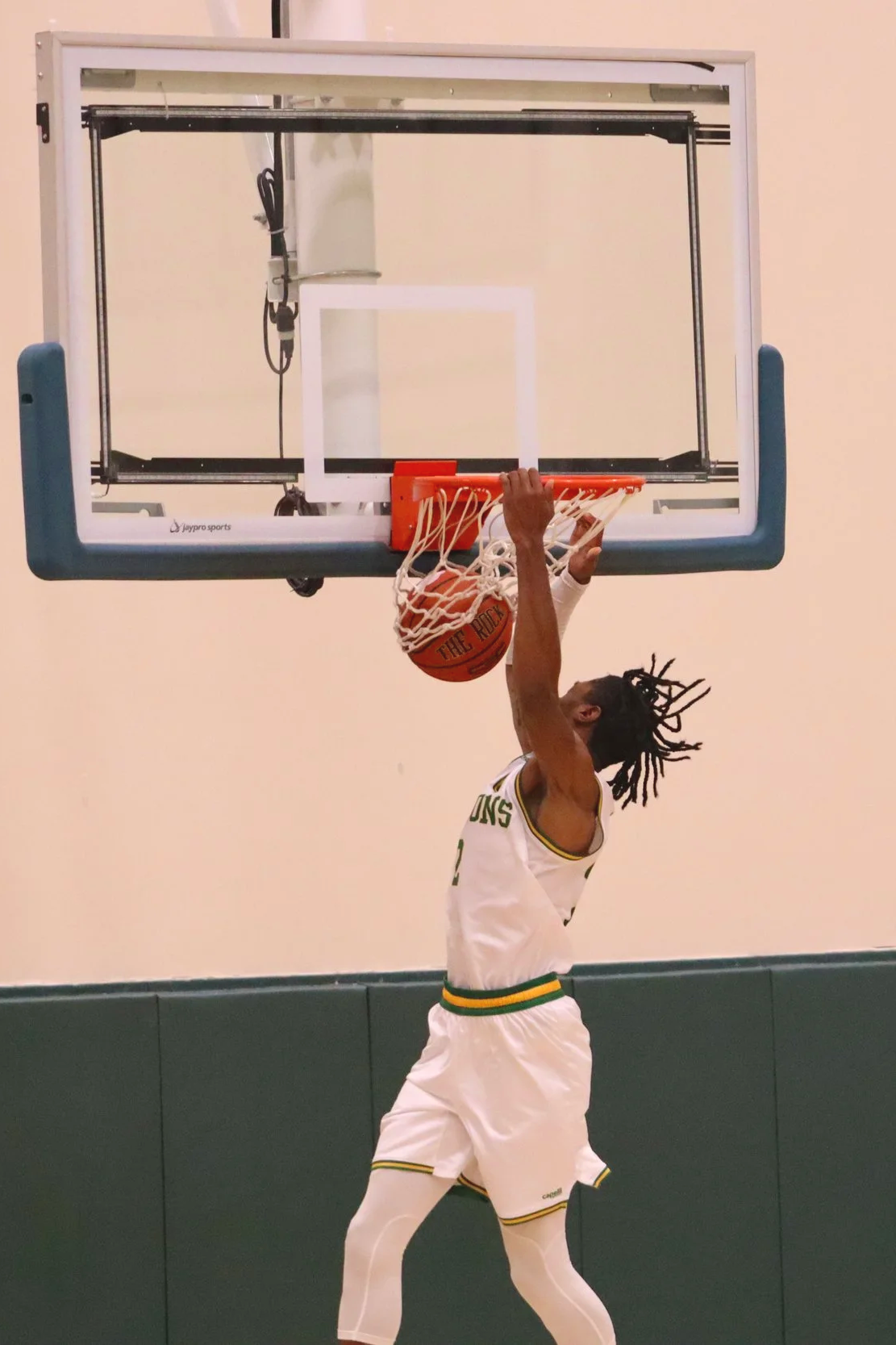 A basketball player in a white uniform with yellow and green accents dunking a basketball into the hoop inside a gym gymnasium.