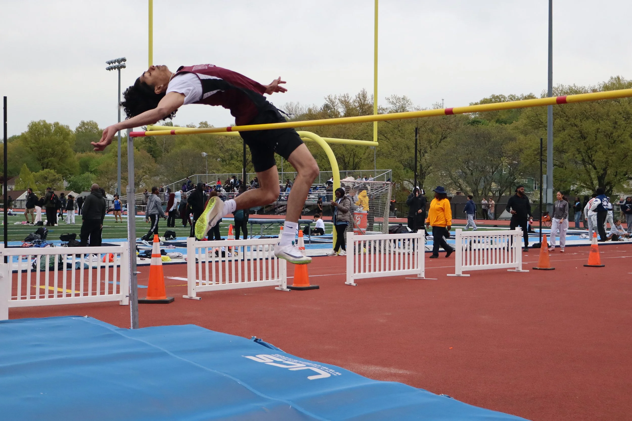 An athlete is mid-air, jumping over a hurdle during a track and field event at an outdoor stadium. There are people in the background watching and participating in the event.
