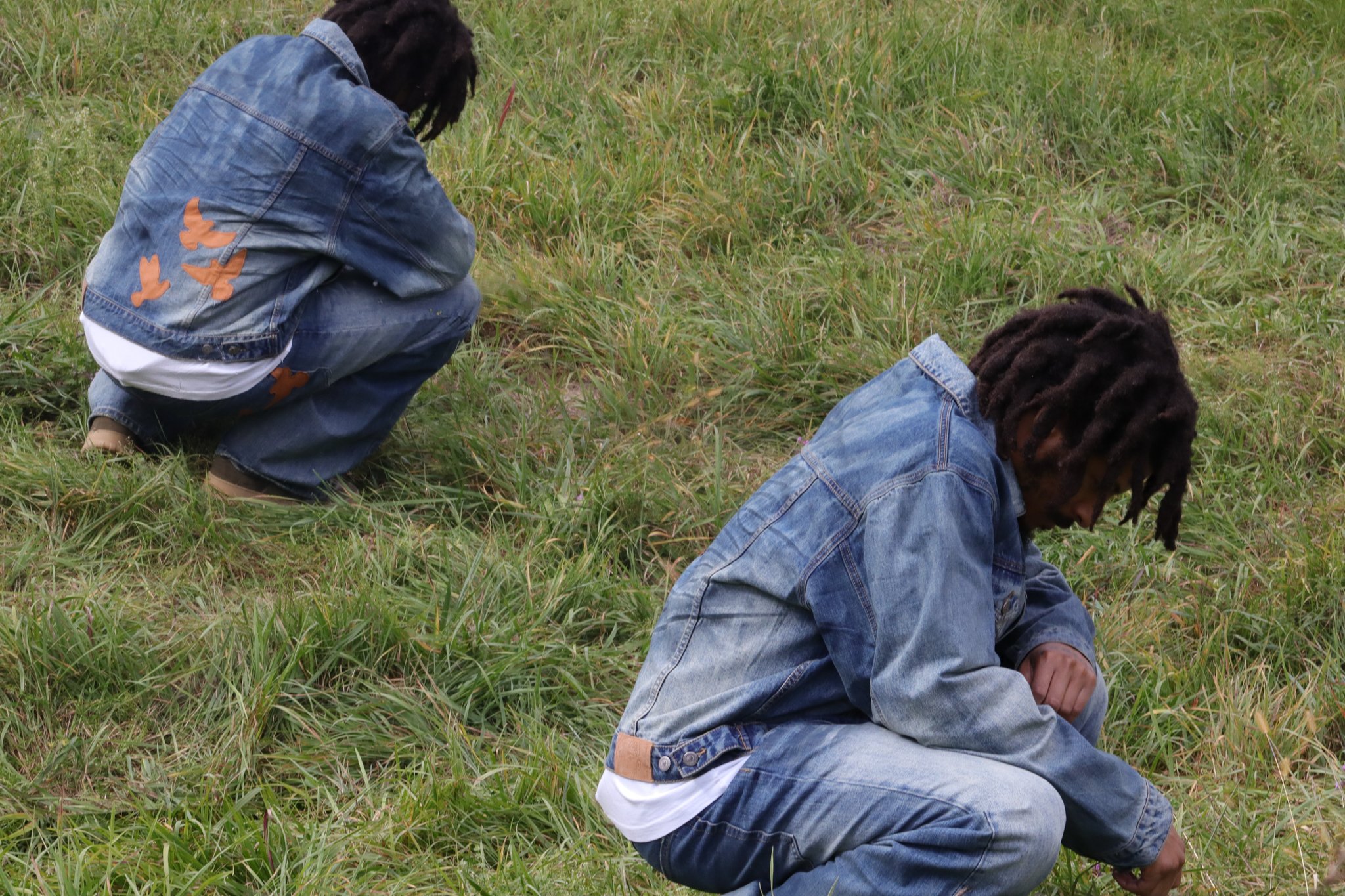 Reflection of Singer with dreadlocks wearing denim jackets, one crouching and one sitting, in a grassy field.