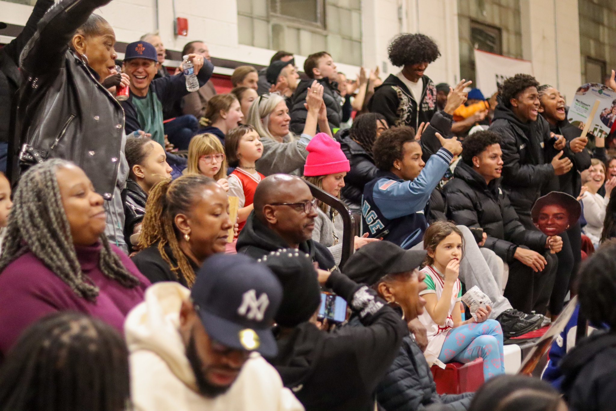 A large group of diverse people, including children and adults, sitting and standing in an indoor stadium, watching and reacting to an event with smiles and expressions of enjoyment.