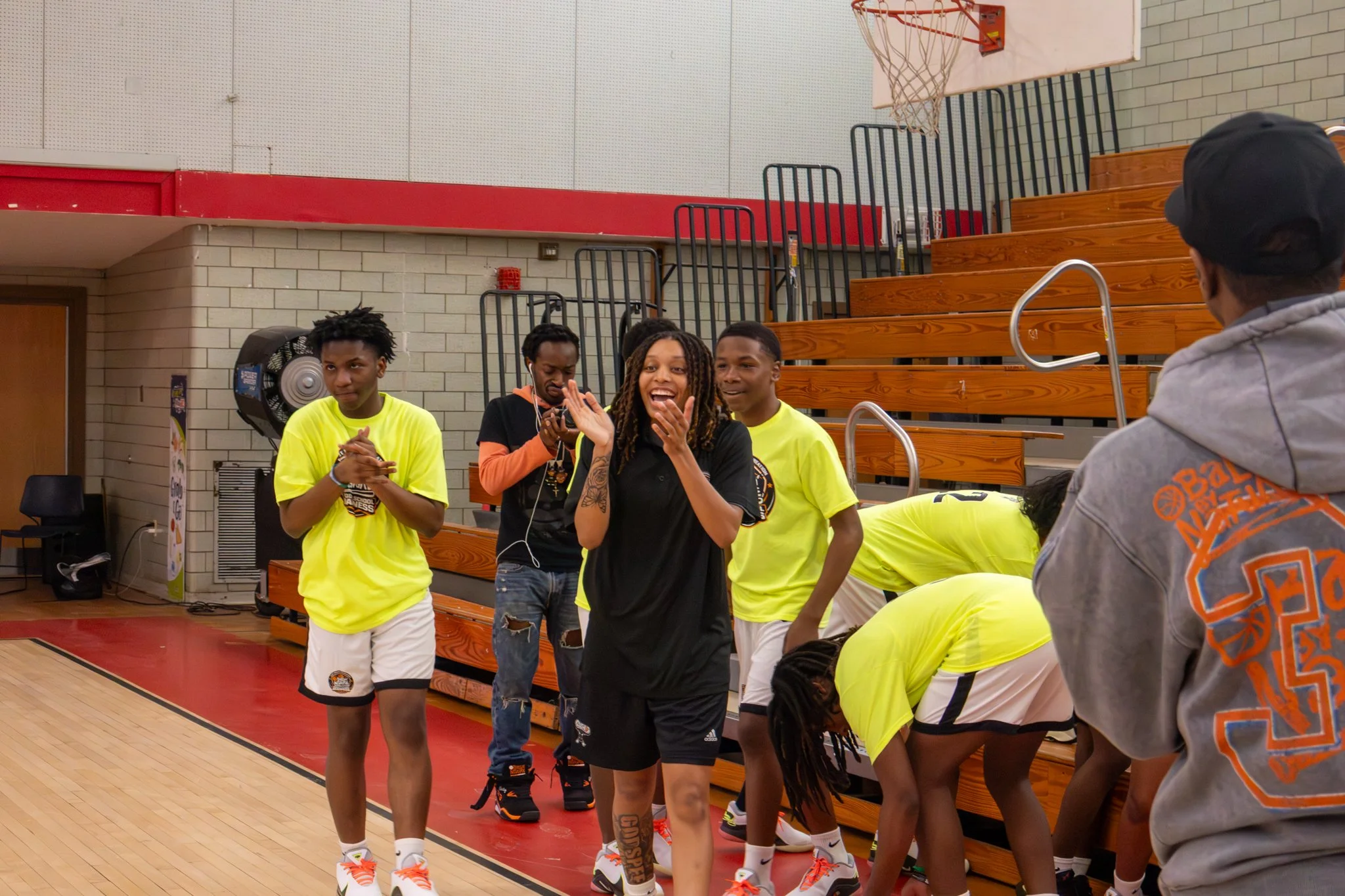 Group of basketball players in a gymnasium, some standing and some kneeling, with smiling and talking among themselves. The players are dressed in yellow and black athletic uniforms, and one person in a gray hoodie is facing away from the camera.
