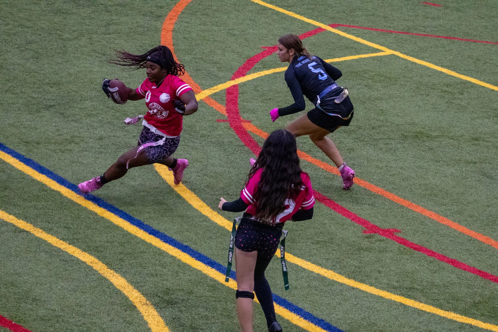 Two women on a synthetic sports field during a game. One woman in pink holds a tackle football, and the other woman in black is running behind her, both concentrating on the game. There are colorful lines on the field.