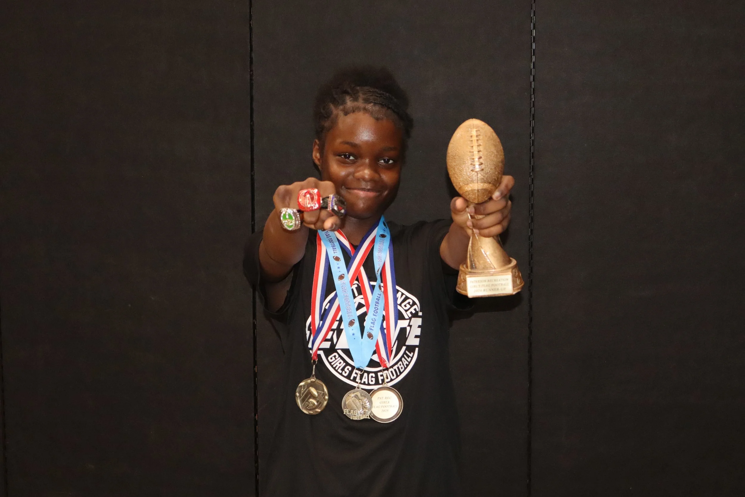 A young girl proudly displaying a trophy and multiple medals, wearing rings and a shirt with text, standing against a black background.