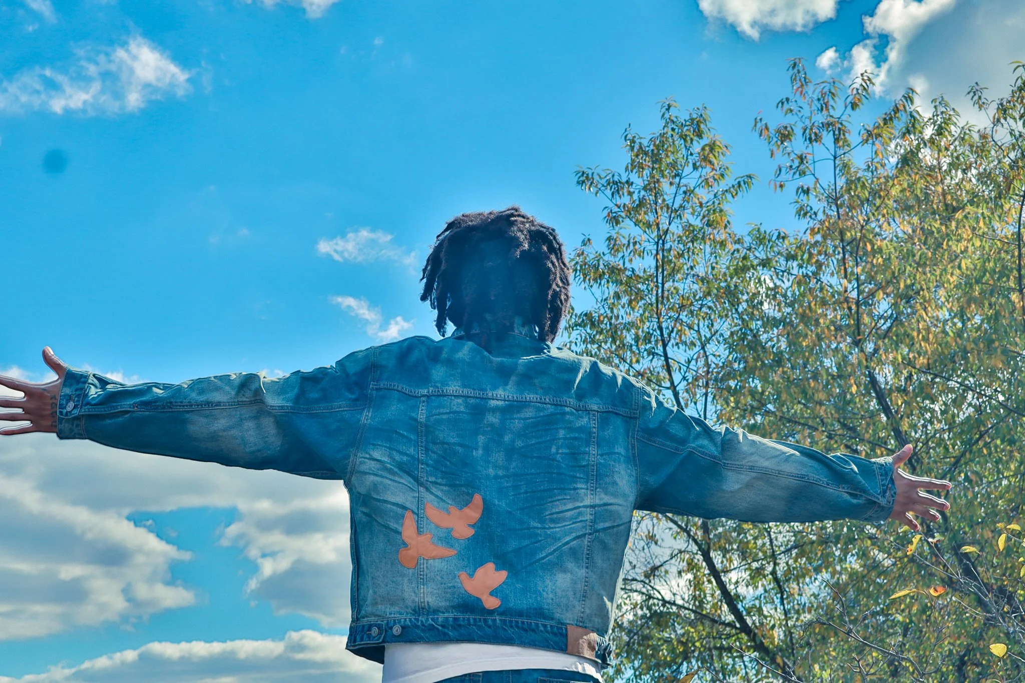 Person with arms outstretched outdoors under a partly cloudy sky, wearing a denim jacket with orange leaf-shaped patches on the back and a butterfly tattoo on the wrist, near trees with green leaves.