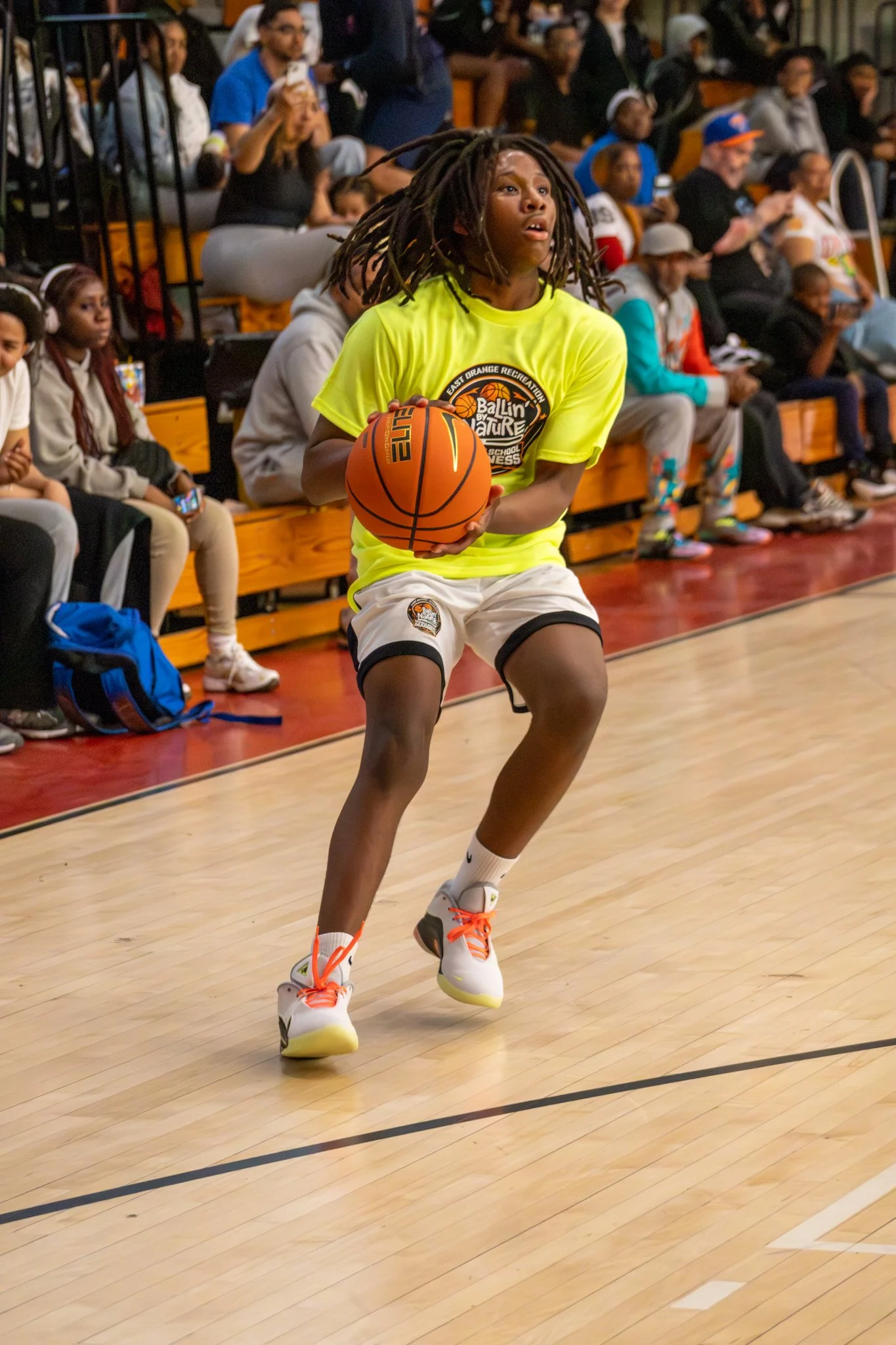A young basketball player with dreadlocks, wearing a bright yellow shirt, white shorts, and white sneakers with orange laces, is holding a basketball on a basketball court. Behind her, there are people seated in the bleachers, including children and 