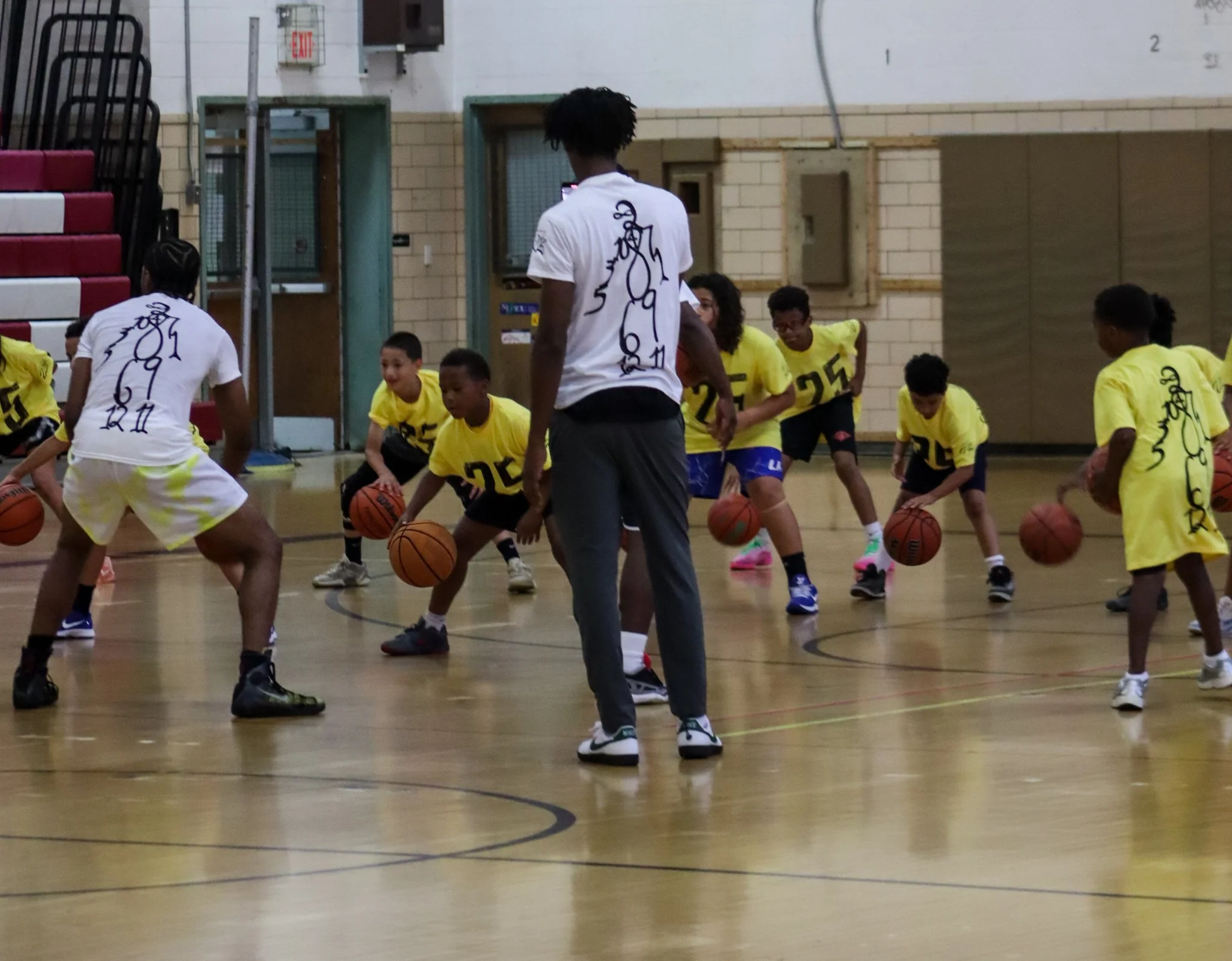 Children in yellow jerseys and a coach in a white T-shirt practicing basketball drills in an indoor gym.