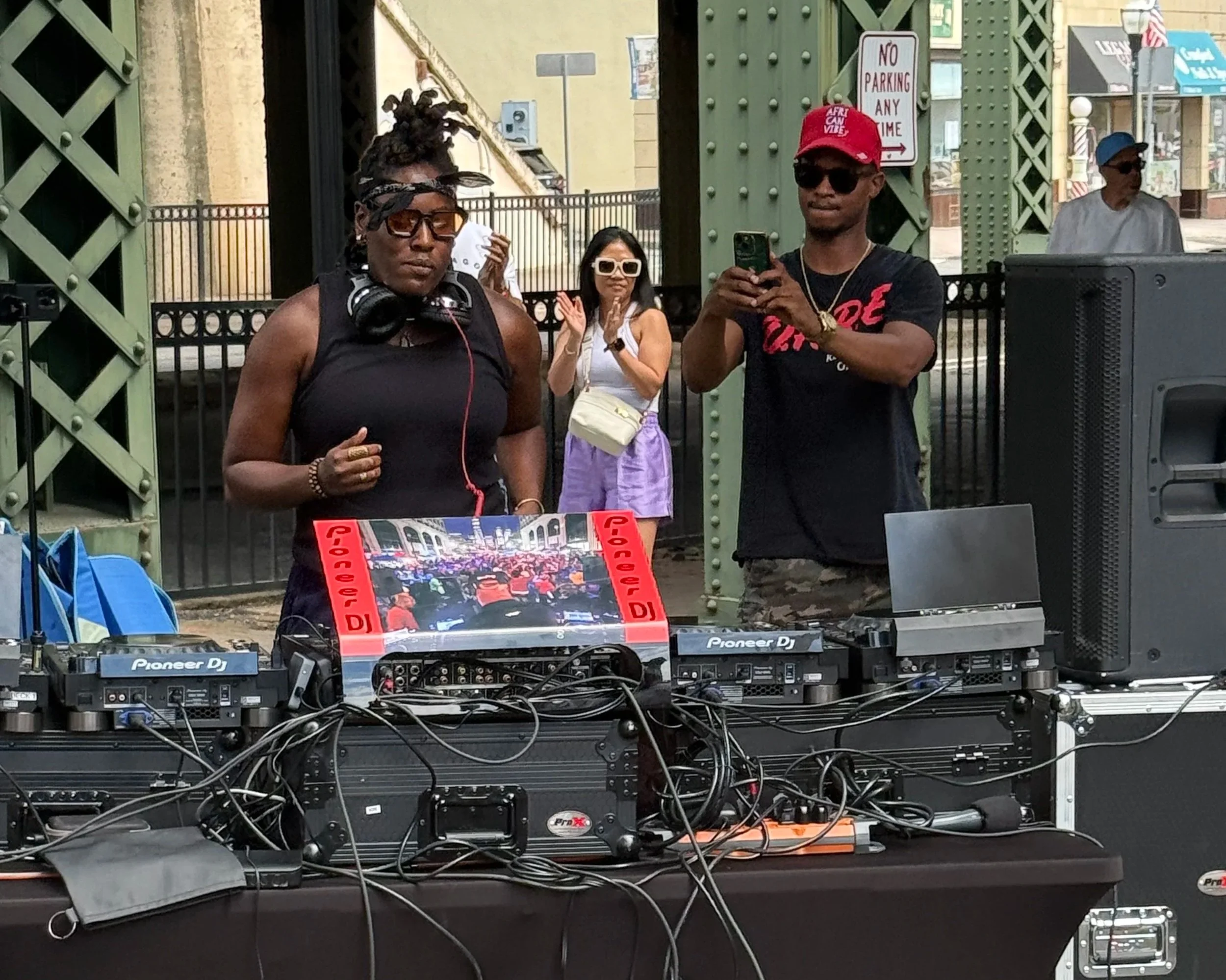 People in front of DJ equipment at an outdoor event, with a woman clapping and a man taking a photo, and a woman in sunglasses in the background.