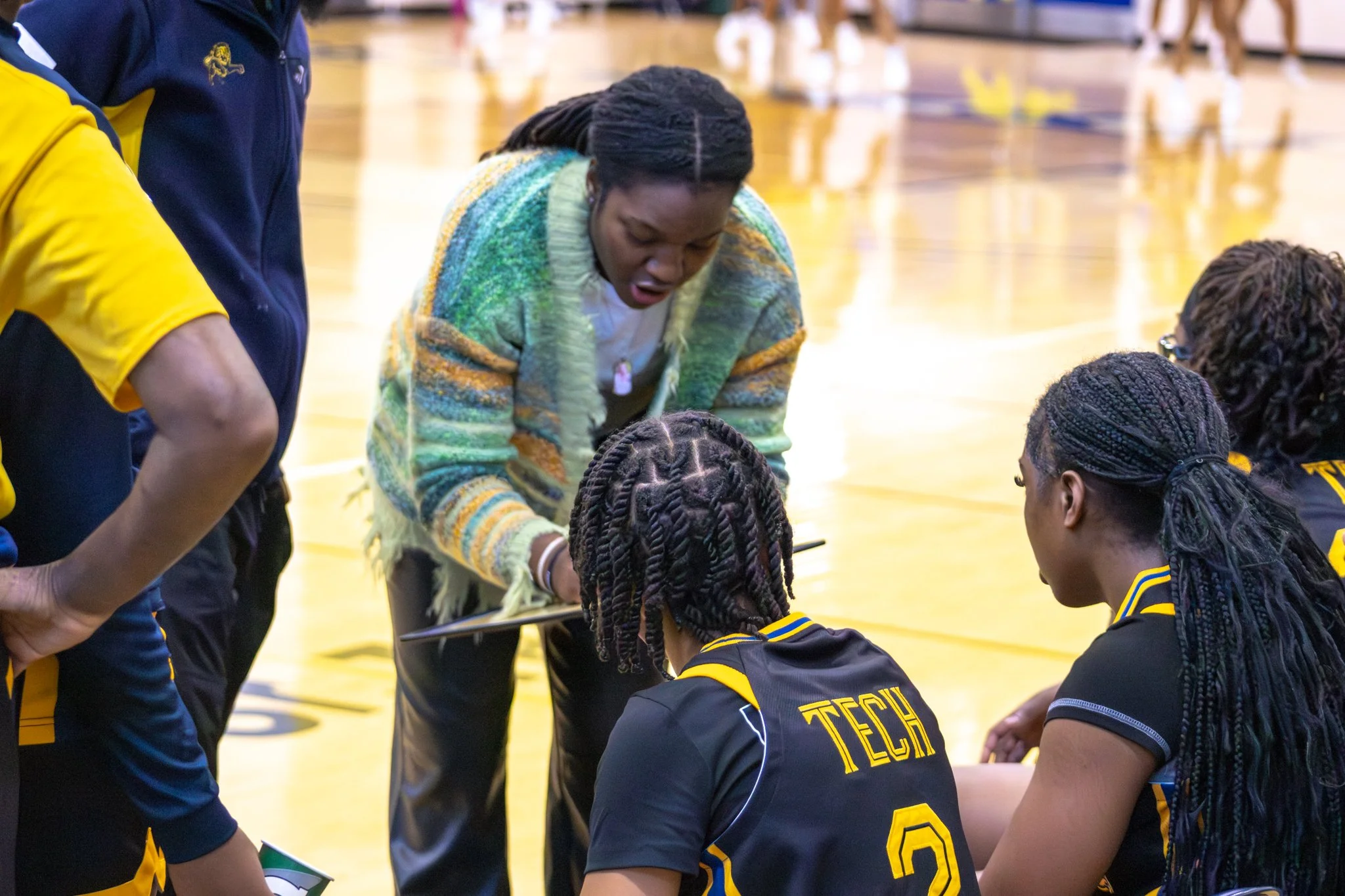 A coach in a colorful sweater is giving instructions to a group of female volleyball players with braided hair wearing black and yellow jerseys in a gymnasium.