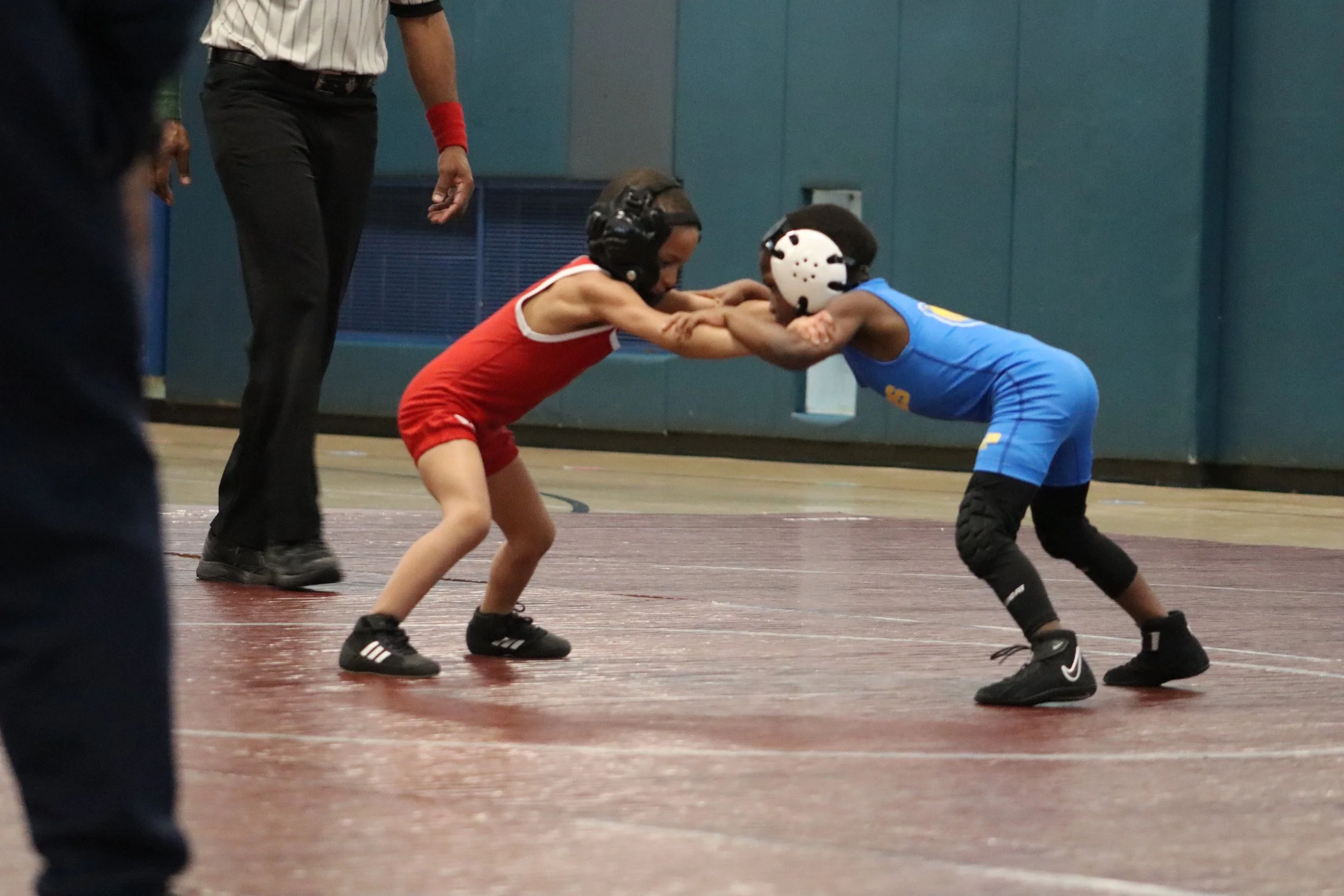 Two young children with protective helmets and athletic gear engaged in a wrestling match on a gymnasium floor, supervised by an adult.