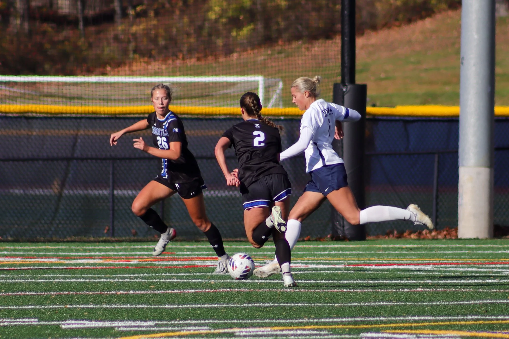 Three female soccer players near the goalpost during a game, with one player in white kicking the ball while two players in black approach
