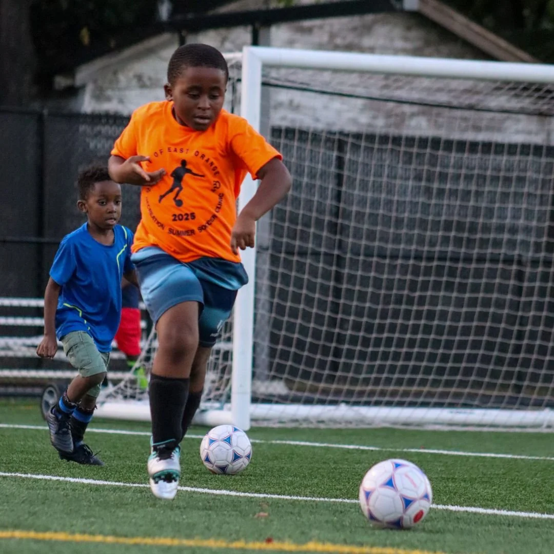 Two young boys playing soccer on a field, one in an orange shirt and the other in a blue shirt, running towards soccer balls near a goalpost.