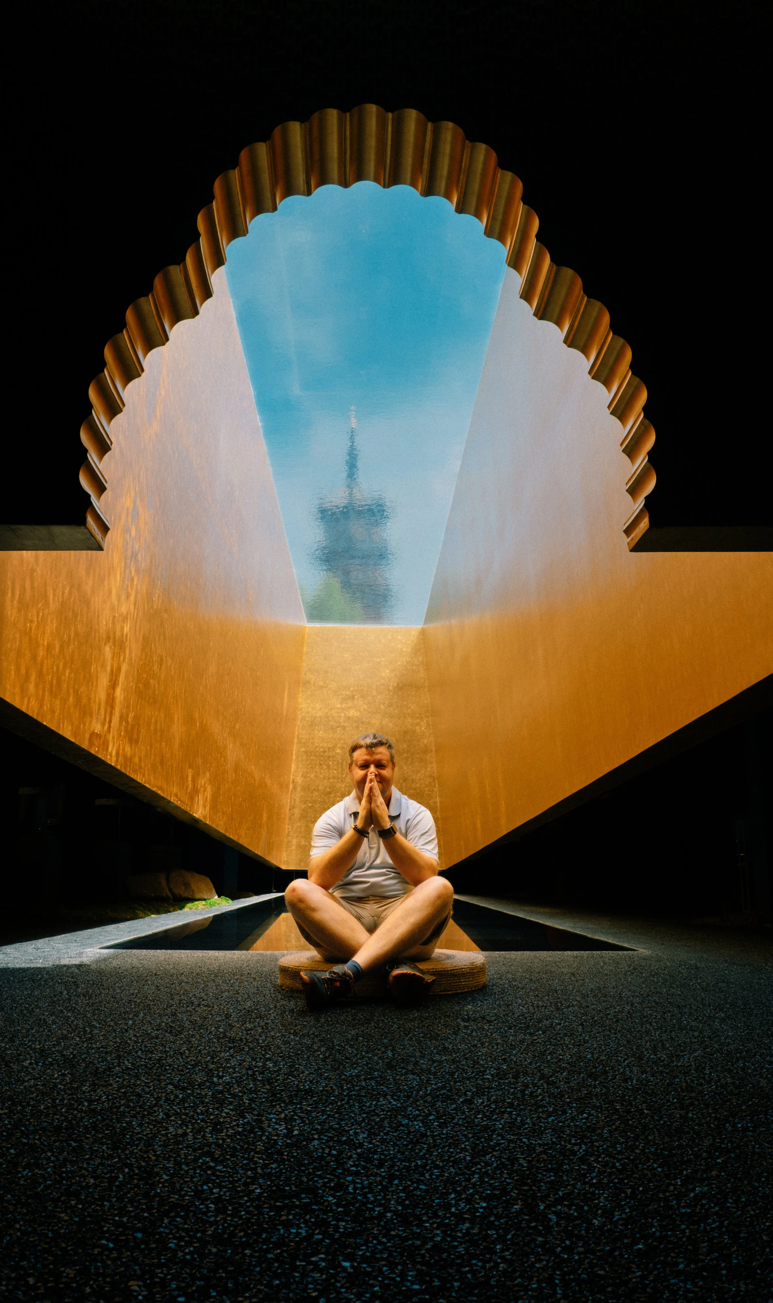 Person sitting cross-legged with hands in prayer position in front of a large, geometric, golden wall with a waterfall and a tower with an observation deck reflected in water above.