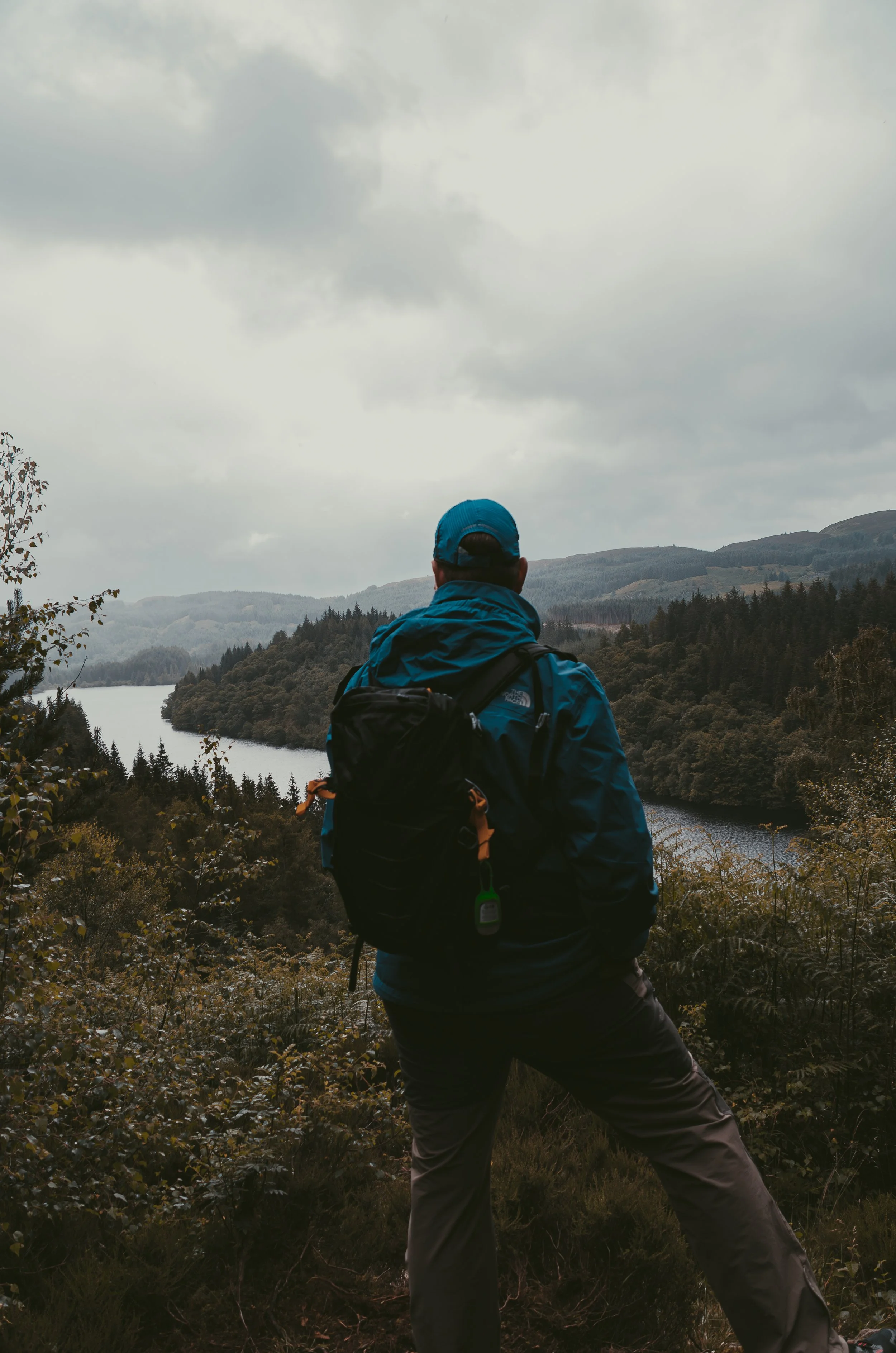 Hiker standing on a trail overlooking a scenic river and forested hills under cloudy sky.