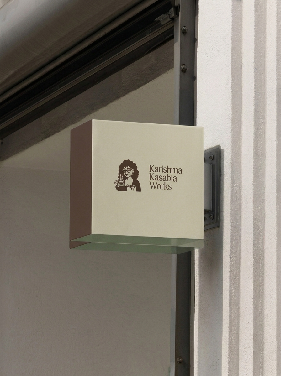 A beige square sign with a cartoon woman with glasses and curly hair holding a coffee mug, alongside the text 'Karisma Kasabia Works', mounted on a wall near a garage door.